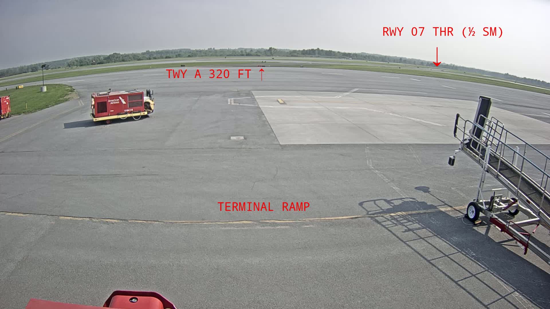 A tarmac on a sunny day shows a baggage tug, a jet bridge, and a runway in the distance.
