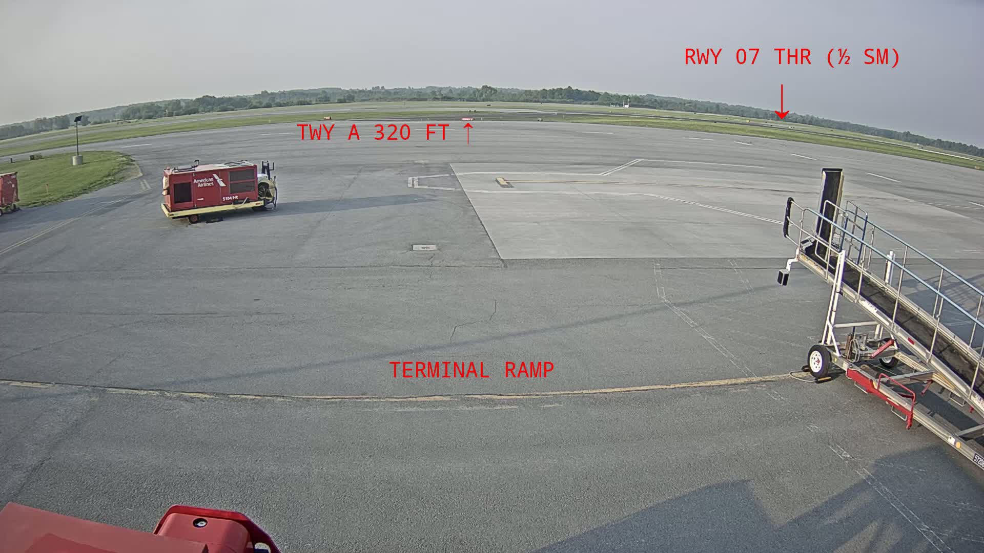 An airport tarmac on a clear day shows a baggage cart, a jet bridge, and a runway in the distance.