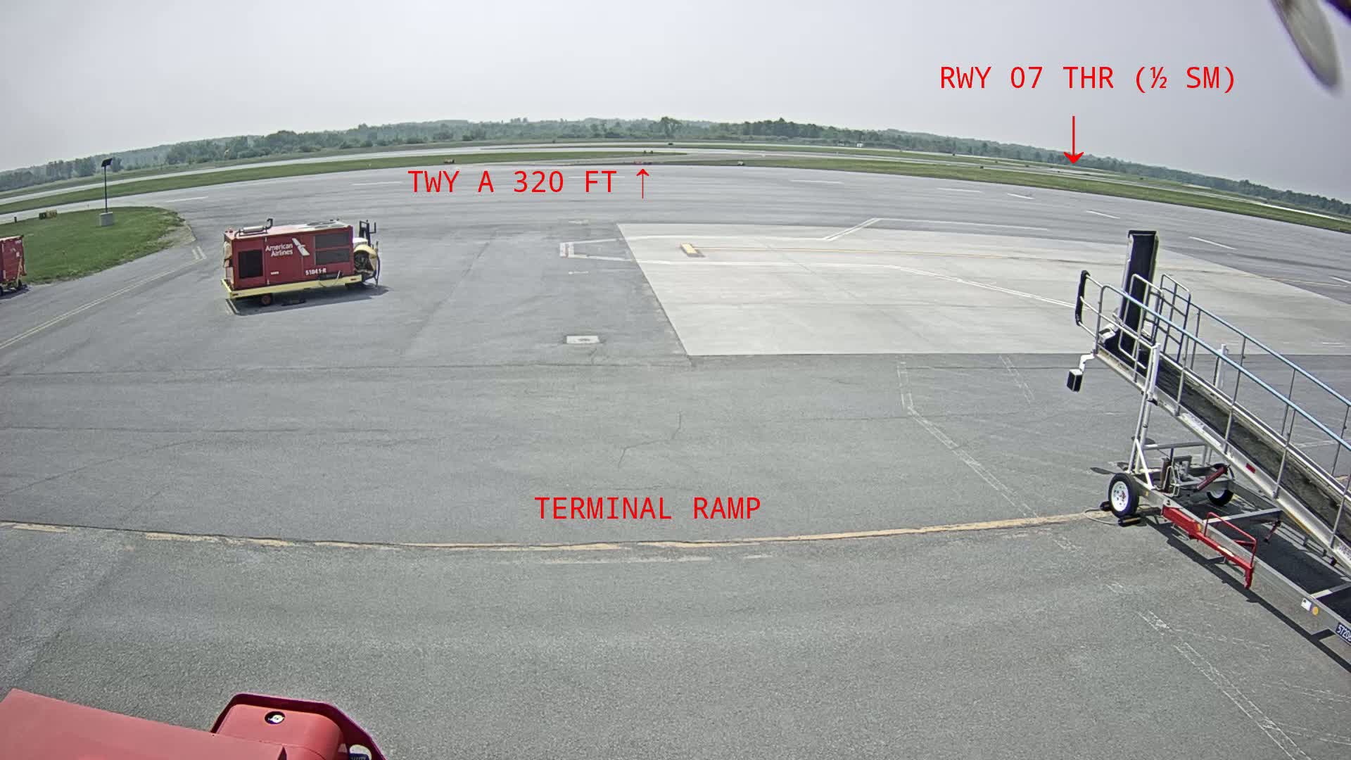 An airport tarmac on a partly sunny day shows a terminal ramp, a runway in the distance, and airport service vehicles.