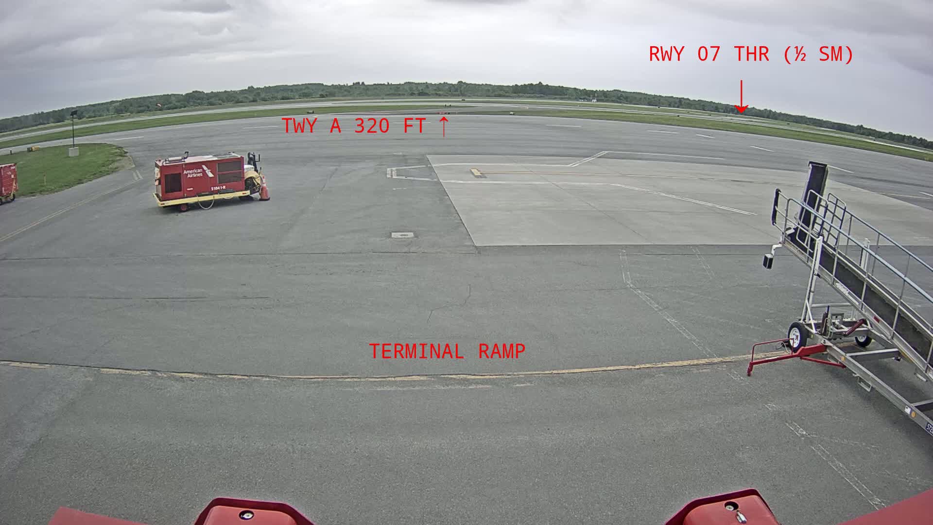 A tarmac view shows a gray airport runway under an overcast sky, with airport service vehicles and a jet bridge present.