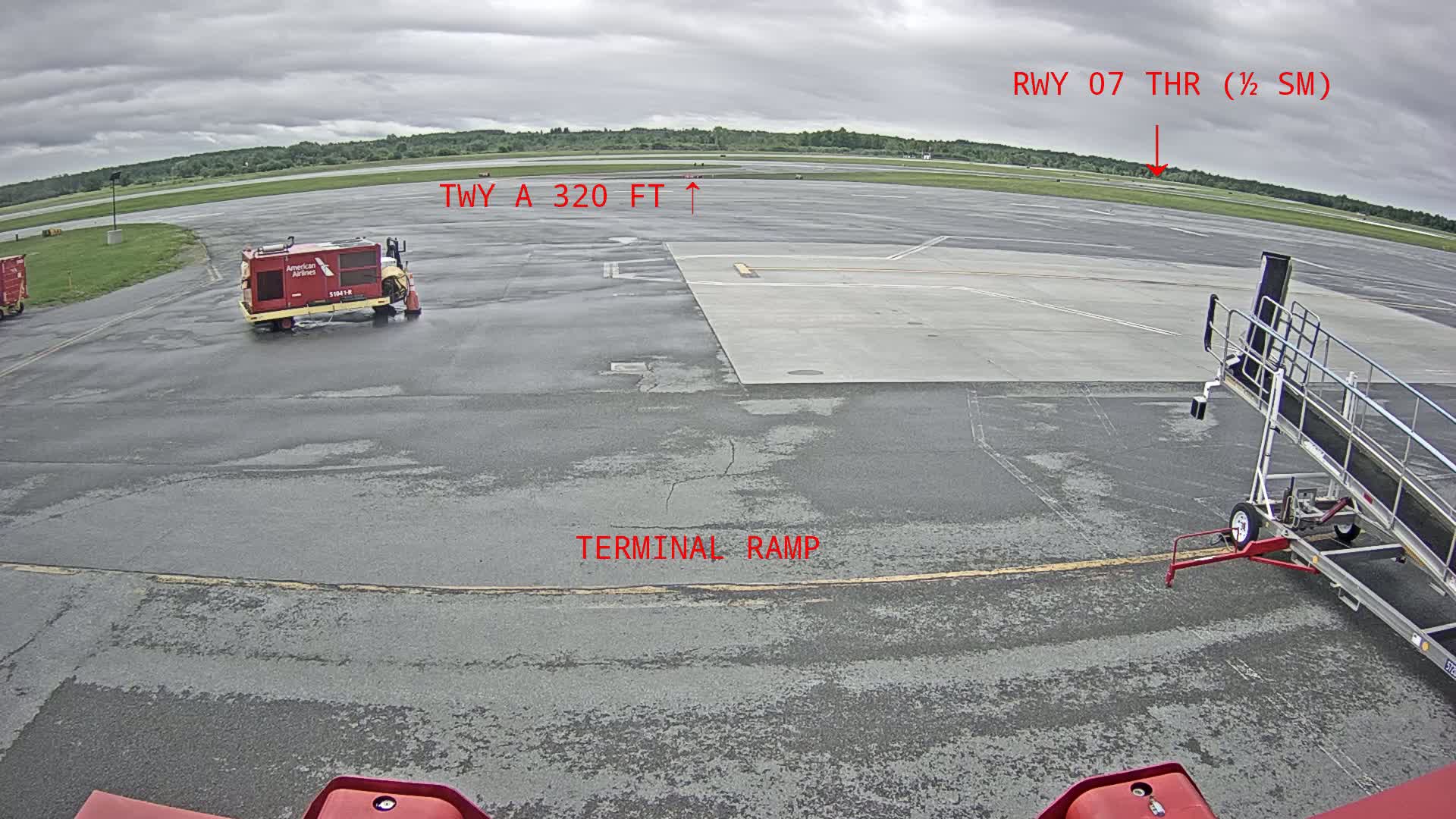 A tarmac under a cloudy sky shows an airport terminal ramp with an American Airlines baggage cart and a jet bridge.