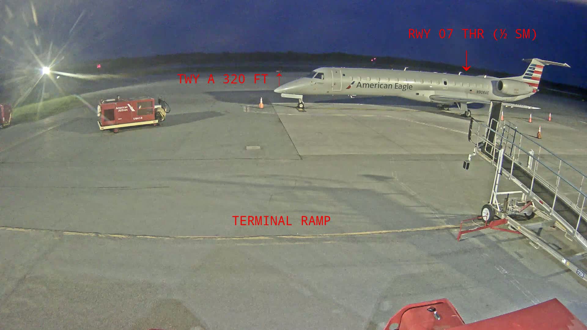 An American Eagle regional jet is parked at an airport gate under a dark, night sky, with airport ground support equipment nearby.