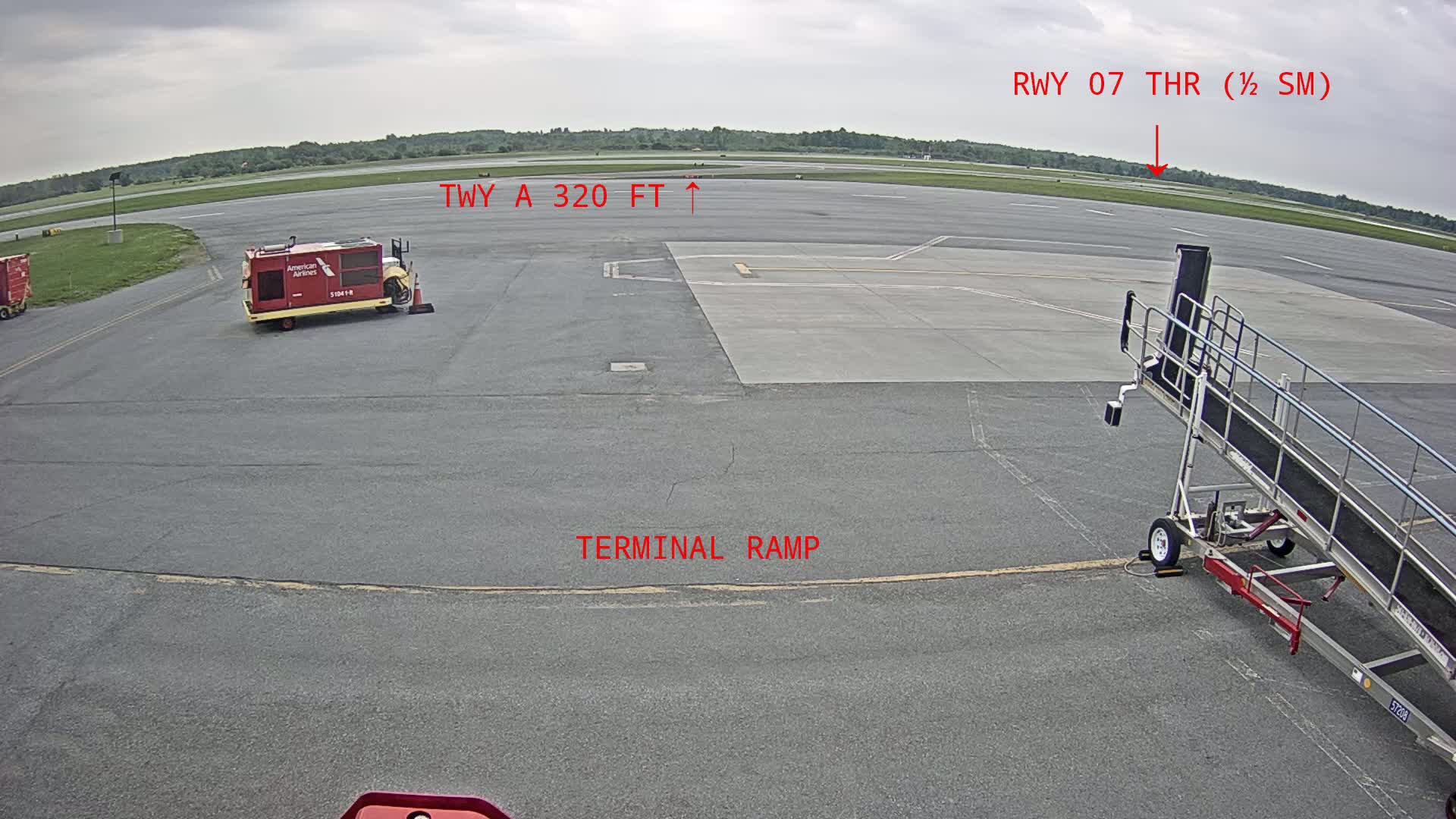 An airport tarmac under overcast skies shows a jet bridge, a baggage cart, and other airport ground support equipment.