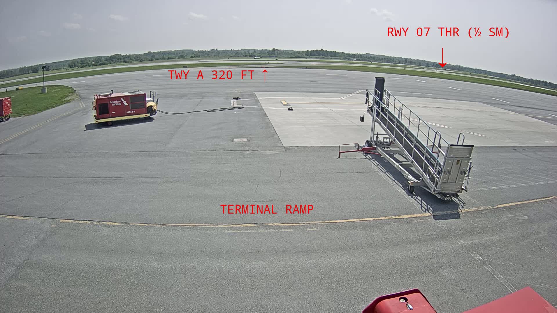 An airport tarmac on a sunny day shows a baggage cart, a jet bridge, and a runway in the distance.