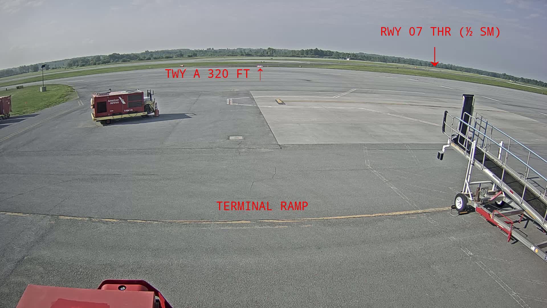 A tarmac at an airport on a partly sunny day shows a baggage cart, a jetway, and a runway in the distance.