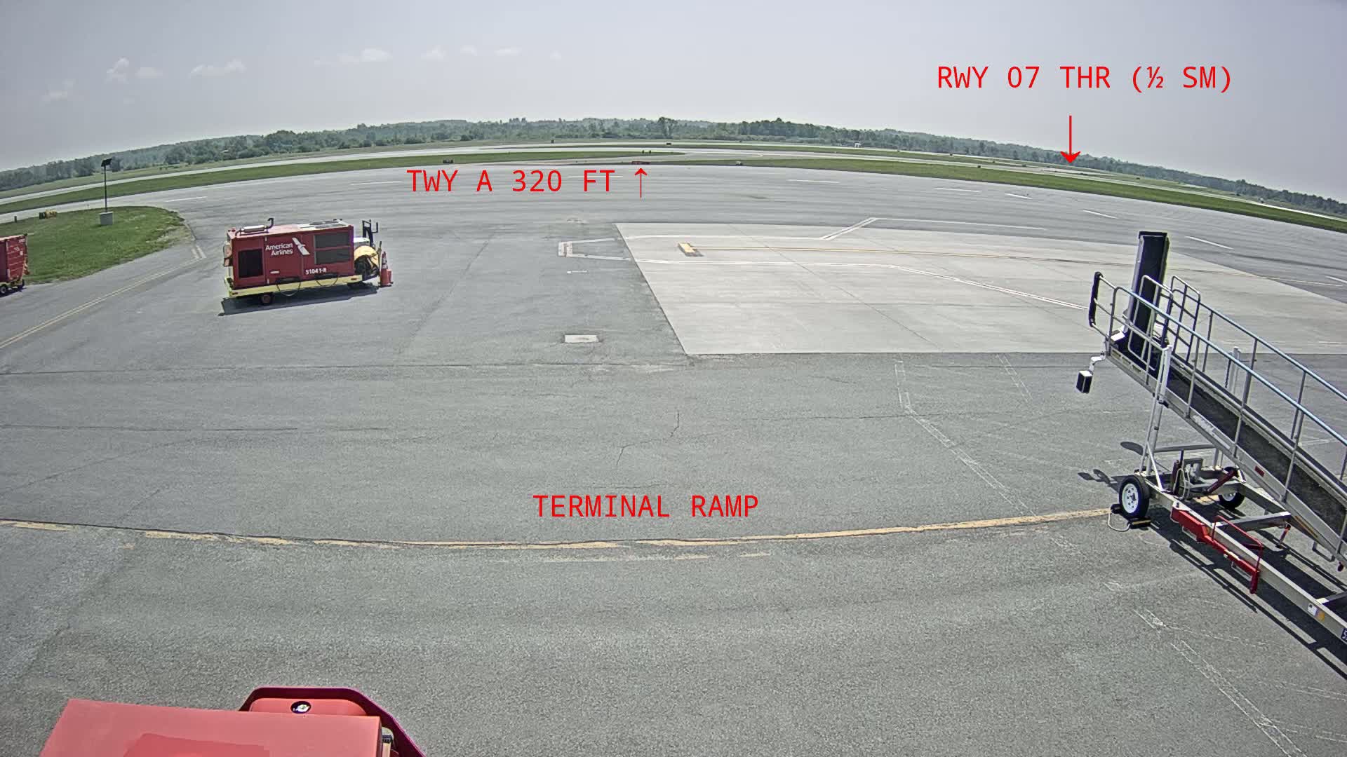An airport tarmac on a sunny day shows a terminal ramp, a baggage cart, and a jet bridge.