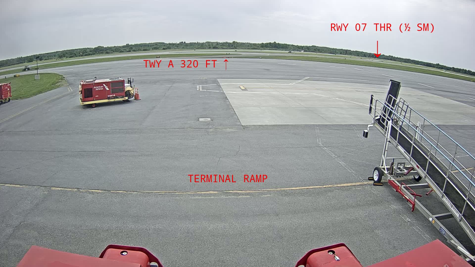 A tarmac at an airport on an overcast day shows an airplane jet bridge, service vehicles, and a runway in the distance.