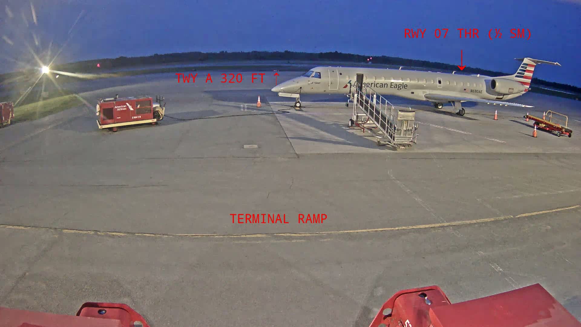An American Eagle regional jet is parked at a terminal ramp at dusk, with ground support equipment nearby.