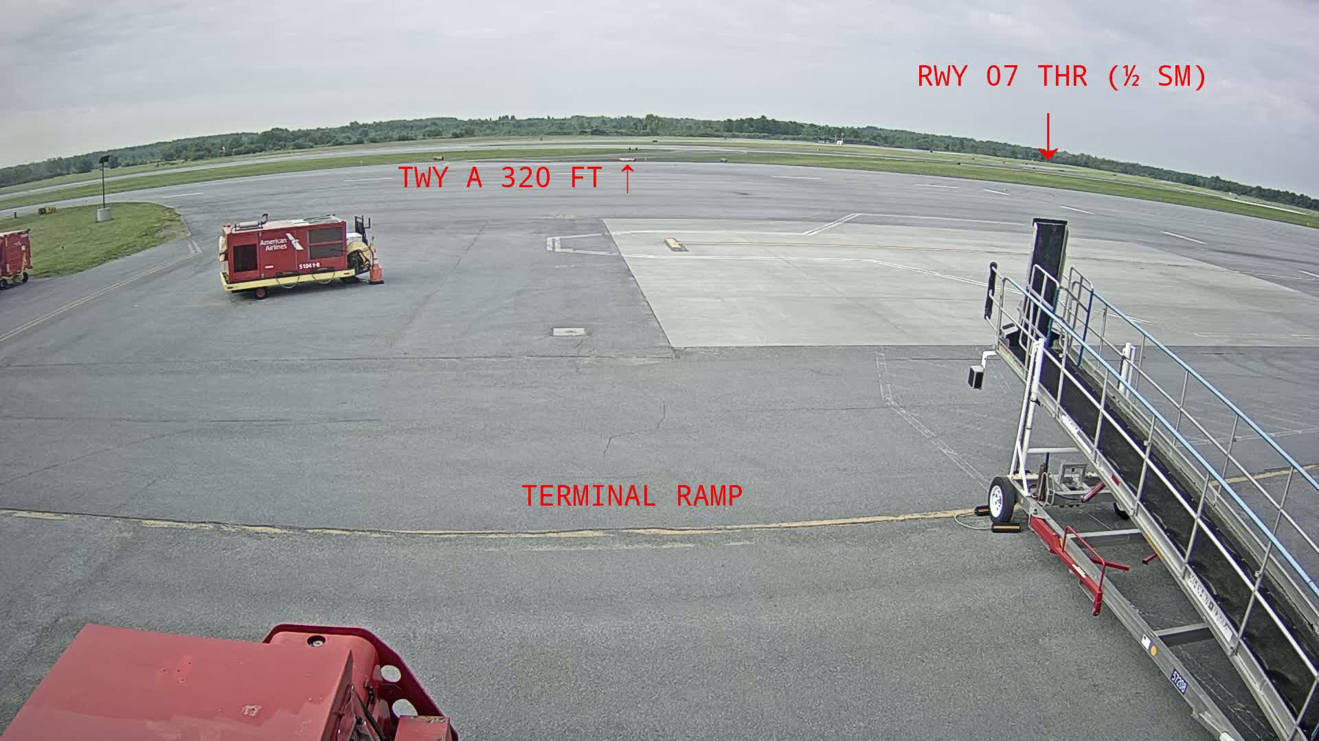 An airport tarmac under overcast skies shows a terminal ramp, a jet bridge, and several airport service vehicles.