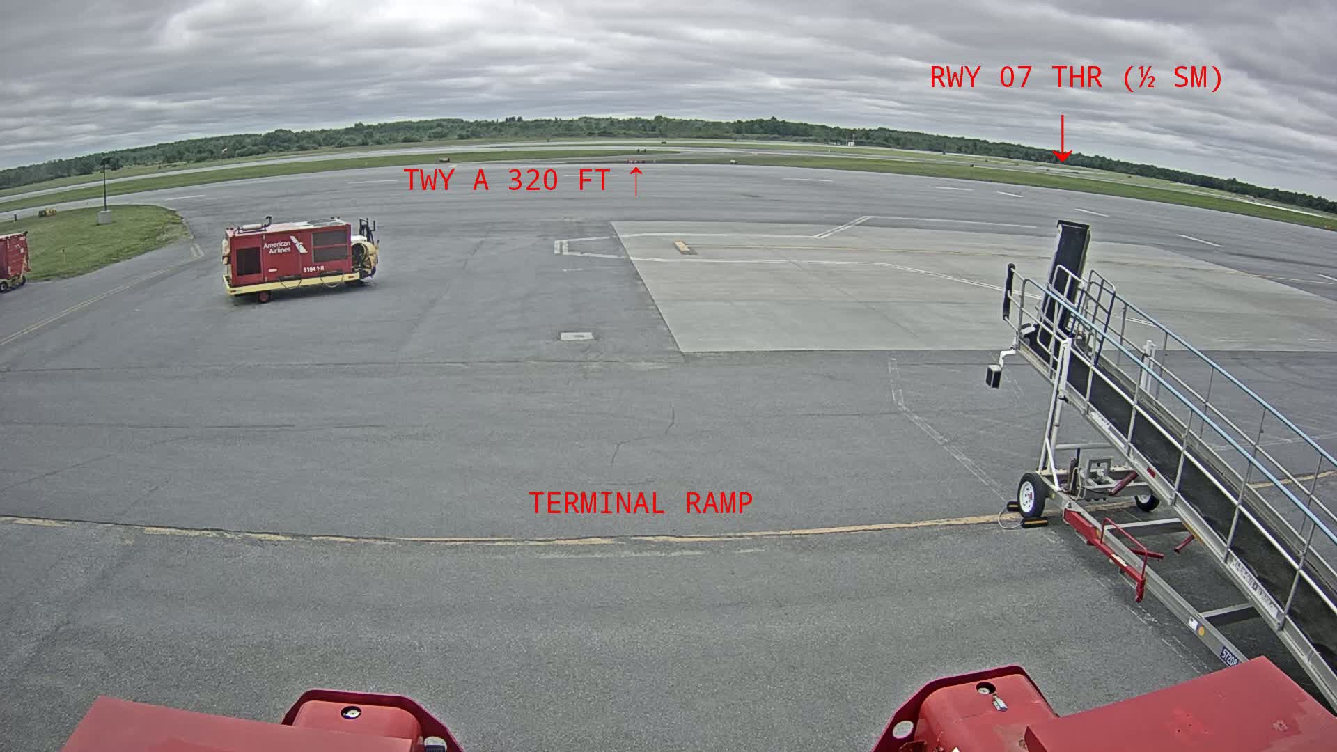 An airport tarmac under overcast skies shows a jet bridge, ground support equipment, and a runway in the distance.