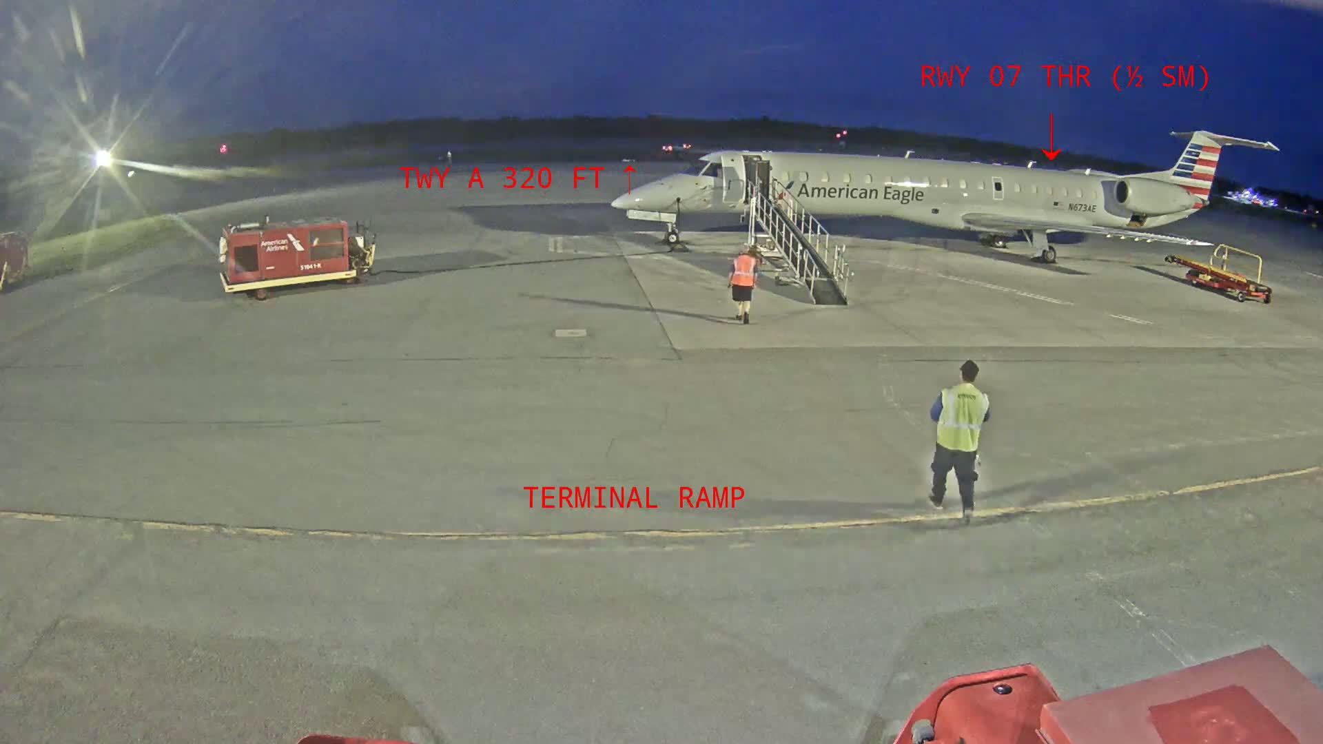 At dusk, an American Eagle airplane is parked at a terminal ramp with airport ground personnel present.