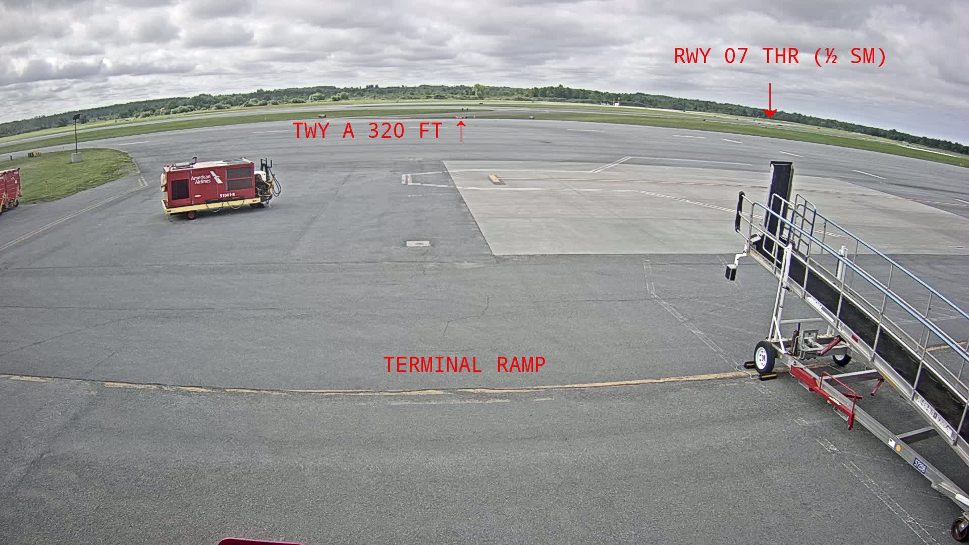 A tarmac under cloudy skies shows airport ground support equipment, a jetway, and a runway in the distance.