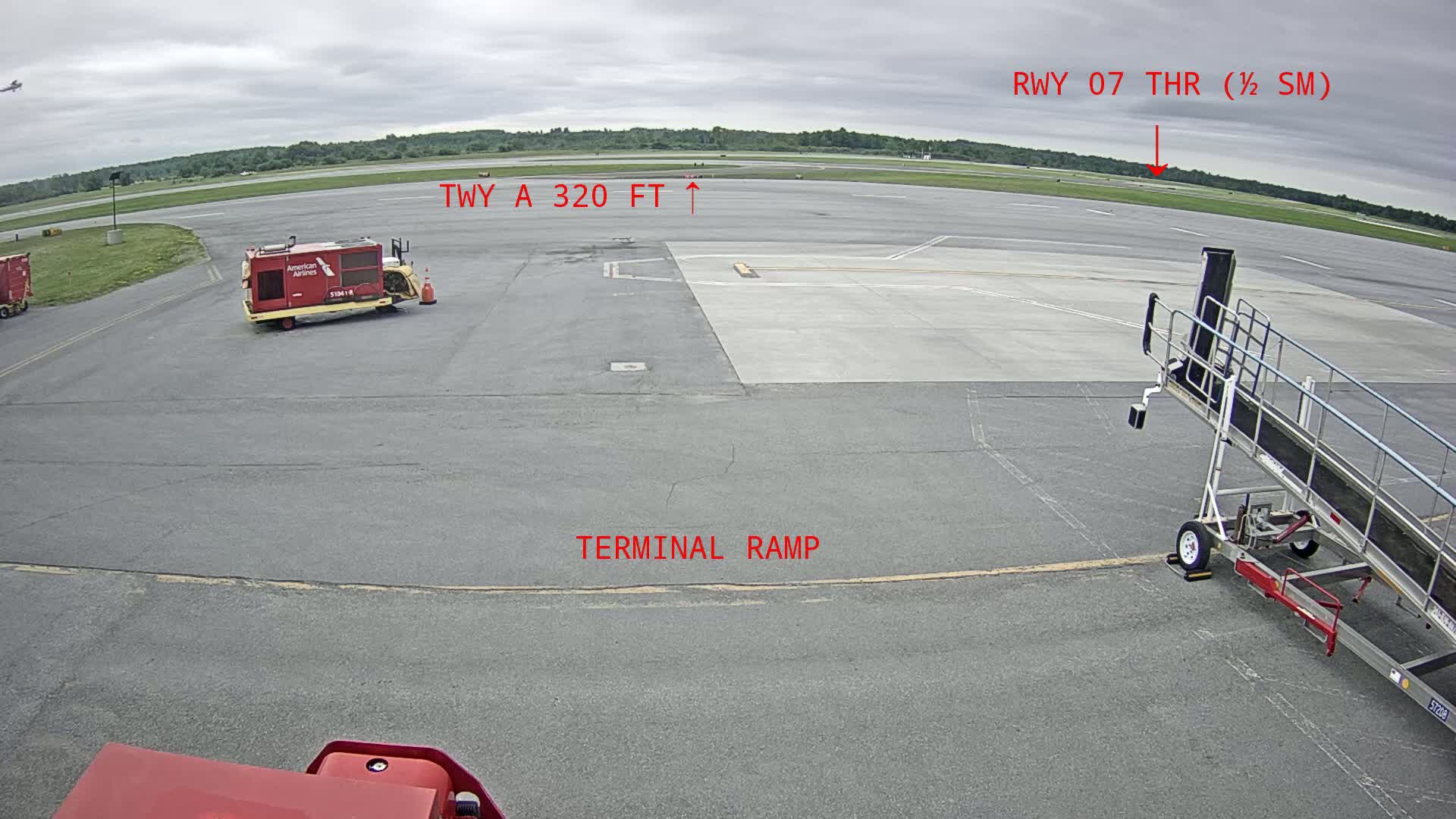 An airport tarmac under an overcast sky shows an airplane servicing vehicle, a jet bridge, and a runway in the distance.