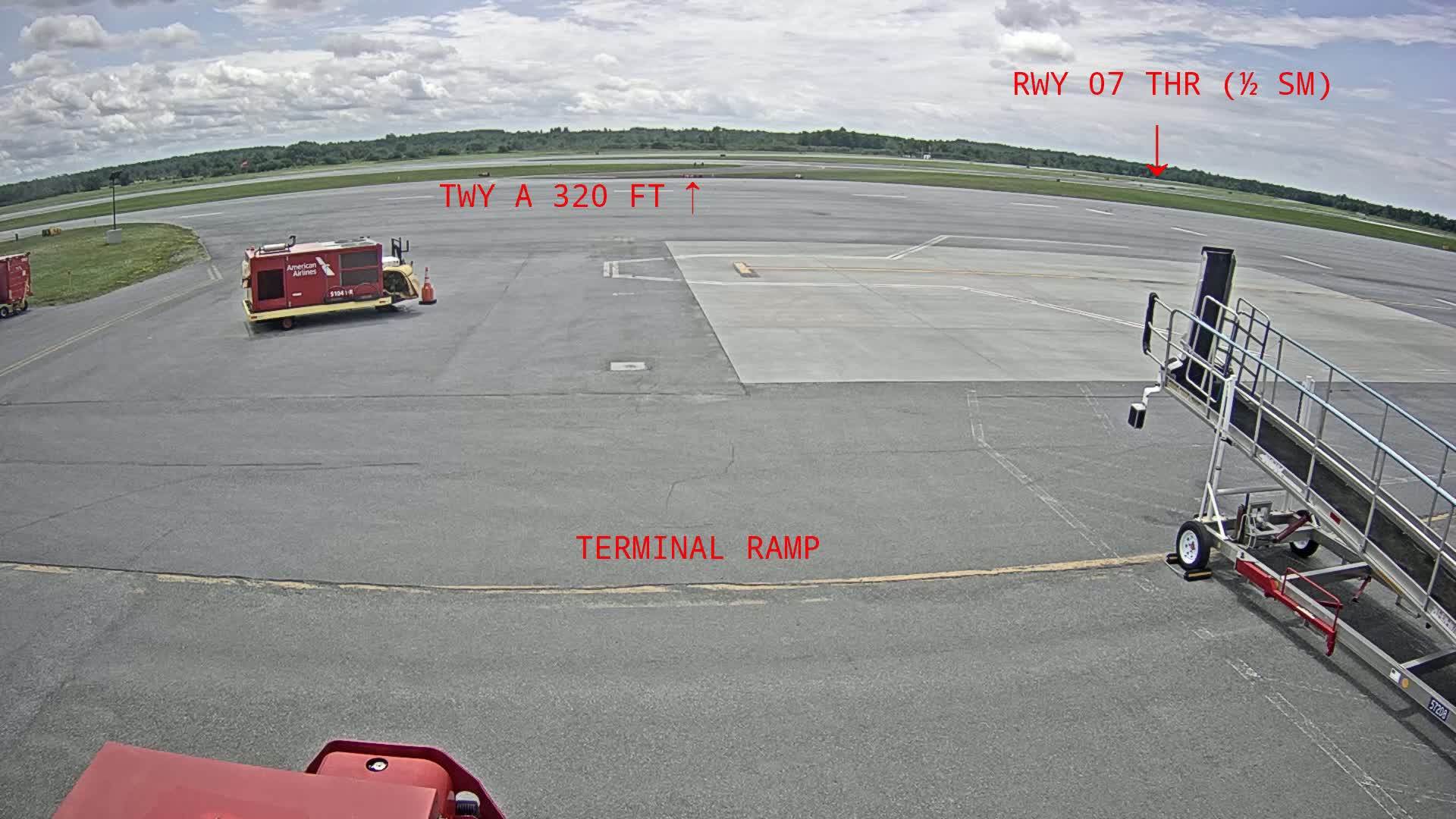 A tarmac at an airport on a partly cloudy day shows a baggage cart, a jetway, and a runway in the distance.