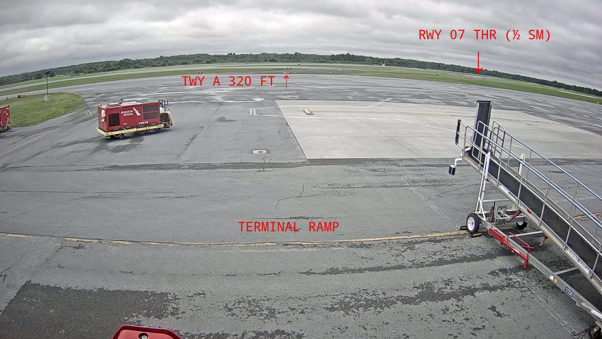 A tarmac under an overcast sky shows an airport terminal ramp, a jet bridge, and airport service vehicles.