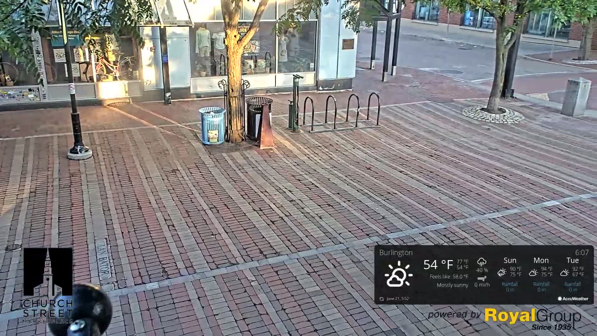 A mostly sunny street scene shows a brick-paved area with a tree, trash cans, bicycle racks, and a lamppost.