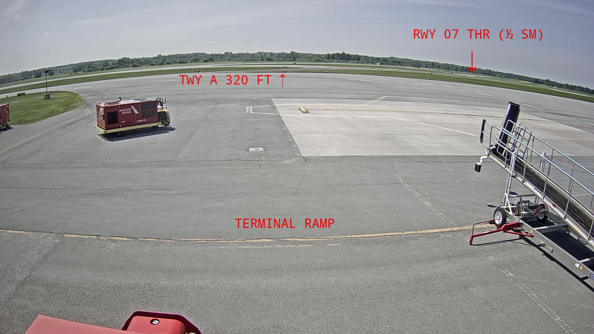 An airport tarmac on a sunny day shows a baggage cart, a jetway, and a runway in the distance.