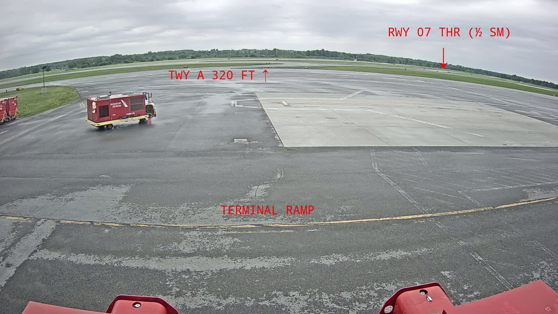 A mostly overcast day at an airport tarmac shows a red baggage cart and other airport equipment, with a runway visible in the distance.