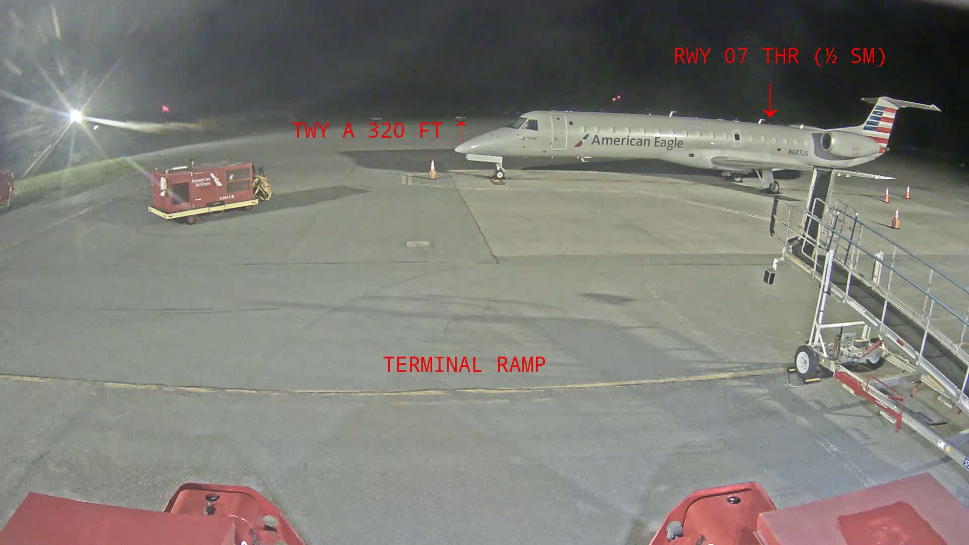 At night, a jet airplane sits on a tarmac near a jet bridge and baggage carts.
