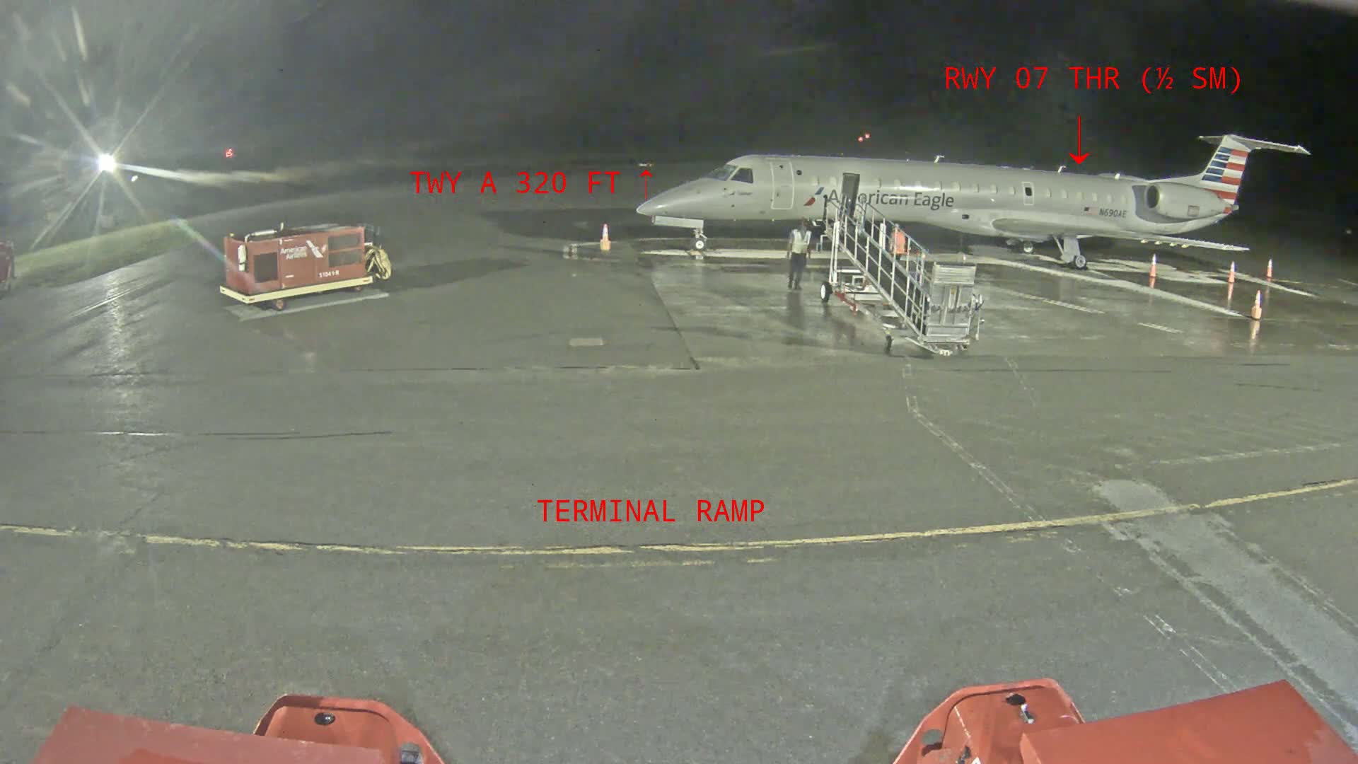 A regional jet is parked at a terminal ramp at night on a wet tarmac with ground support equipment nearby.
