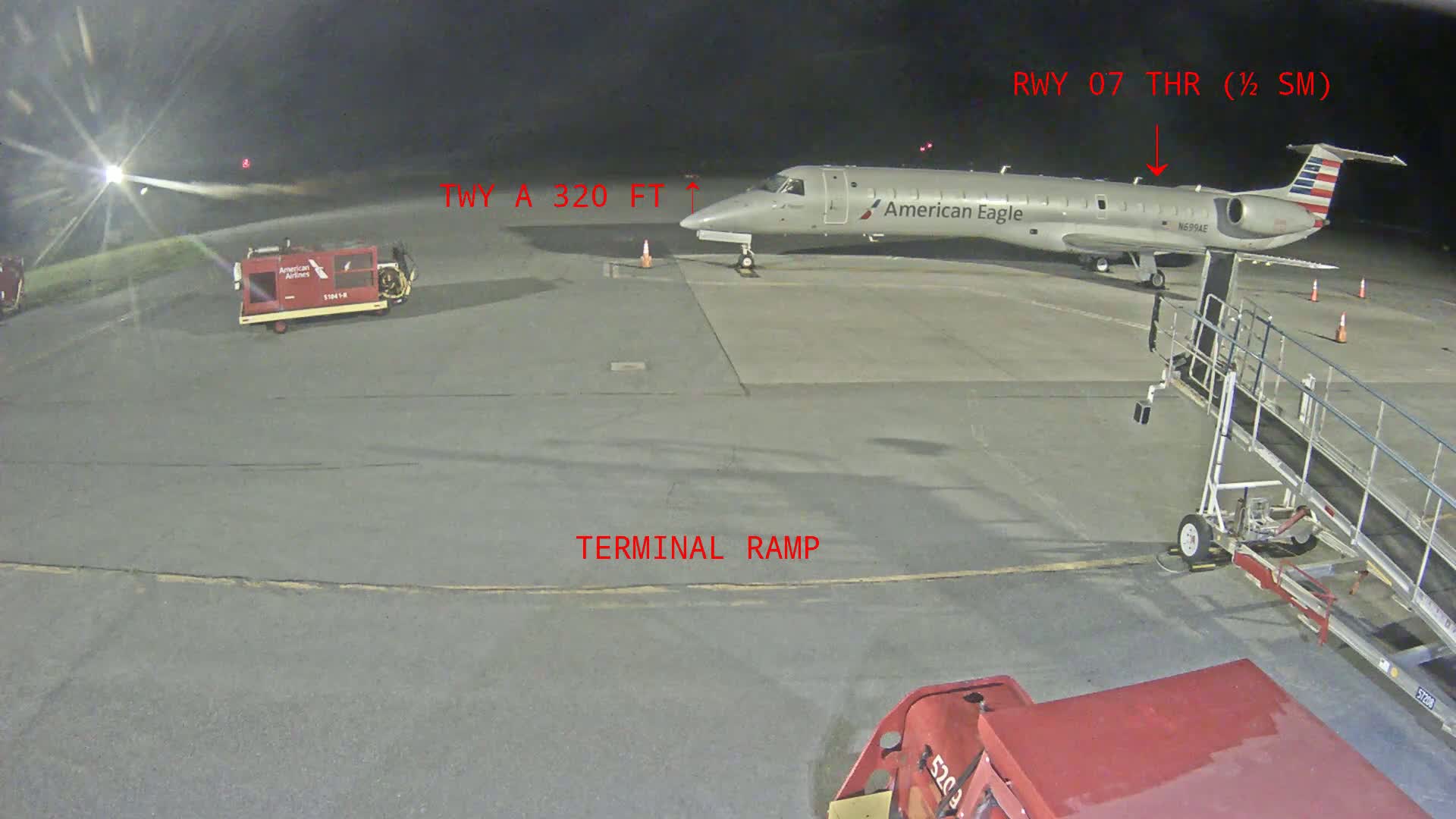 An American Eagle regional jet is parked on a tarmac at night next to a jet bridge and ground support equipment.