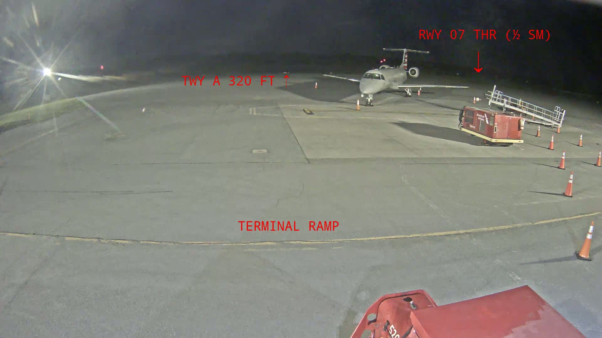 A regional jet is parked on a tarmac at night near a terminal ramp and baggage cart, with airport lighting illuminating the scene.