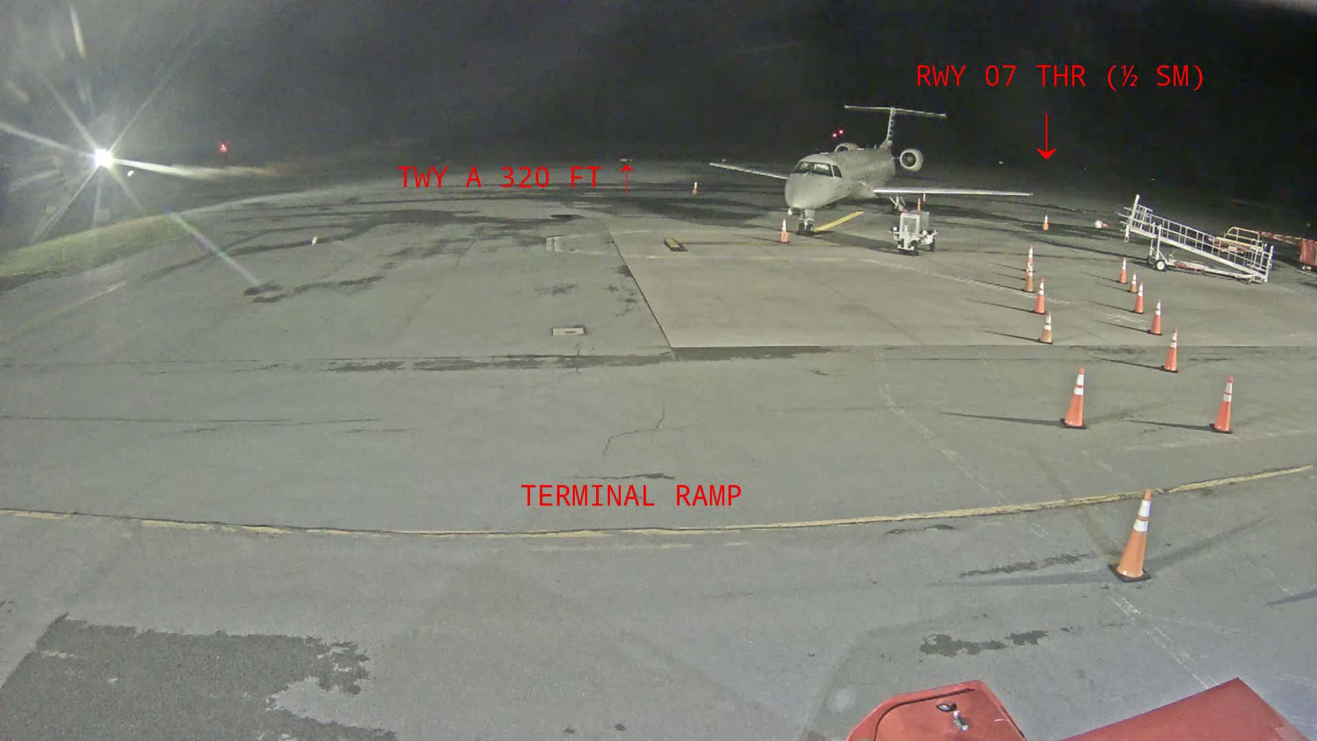 A small passenger plane sits on a dark airport tarmac at night, surrounded by orange traffic cones and airport equipment.