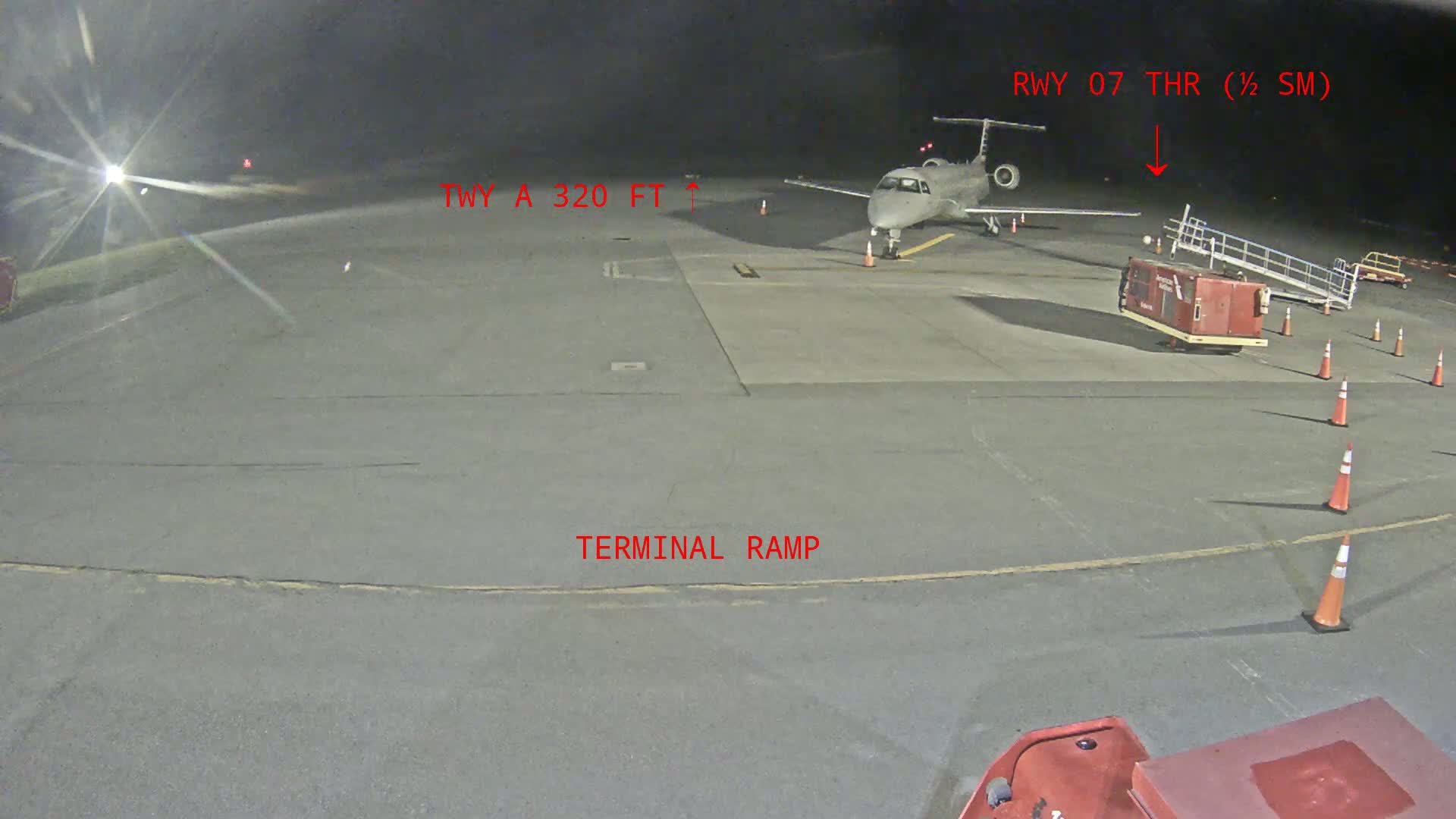 A small passenger plane sits on a tarmac at night, near a baggage cart and several traffic cones.