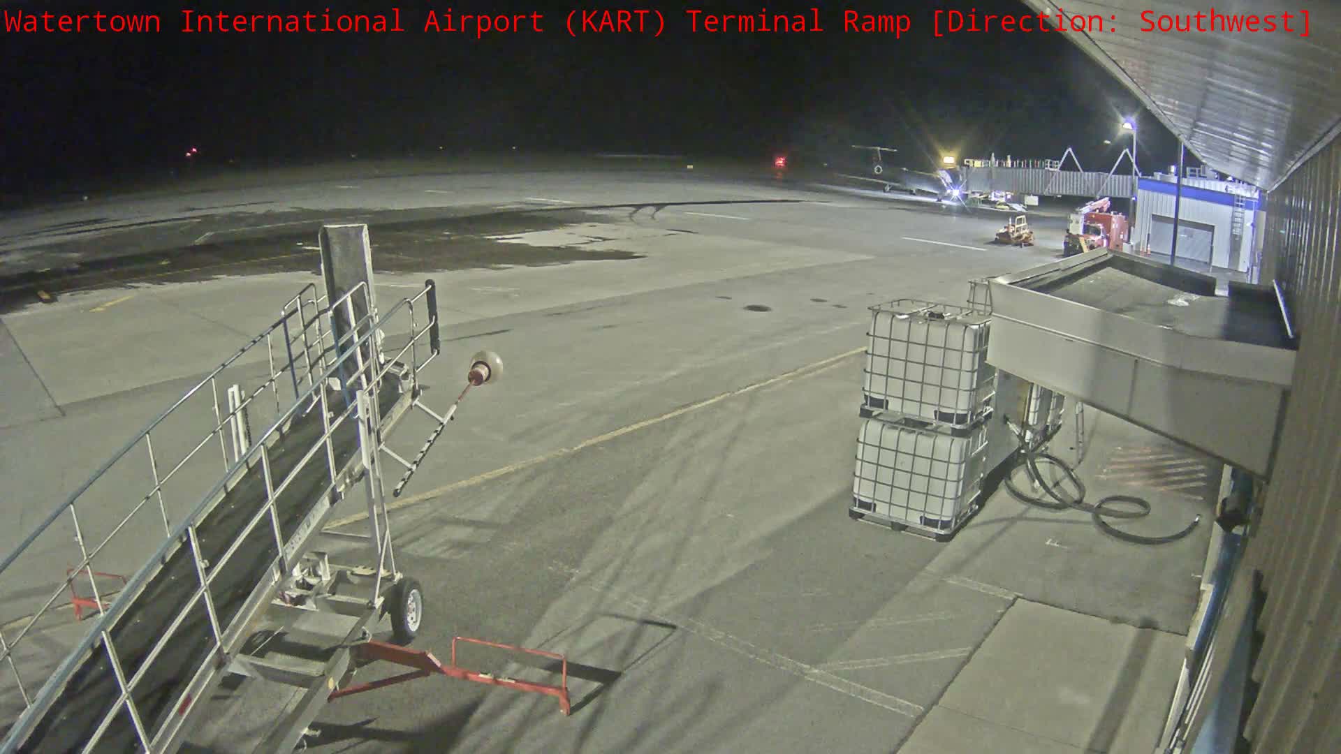A brightly lit airport terminal ramp is seen at night, featuring a parked aircraft connected to a jet bridge, various ground support equipment, and sections of the terminal building, all under clear skies.