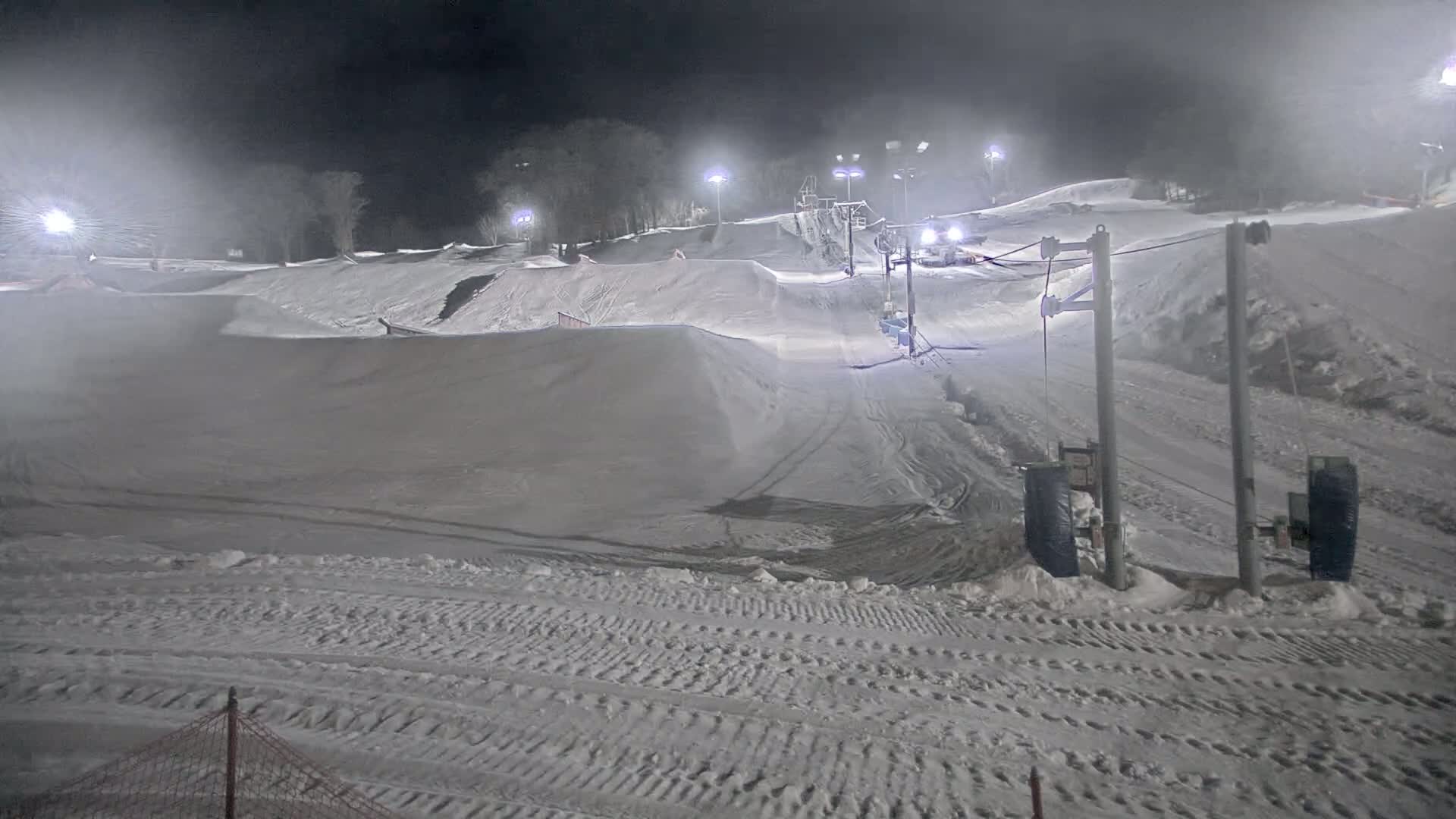 A snow-covered terrain park at night, illuminated by lights, with a ski lift and snowy hills under foggy conditions.