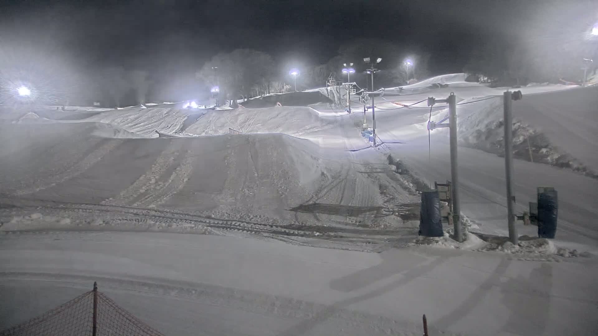 A snow-covered ski slope at night, lit by artificial lights, with a surface lift visible and some snow-making equipment.