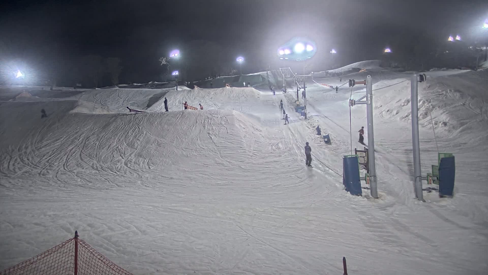 A snow-covered ski slope at night is lit by floodlights, with several skiers and snowboarders using a surface lift.