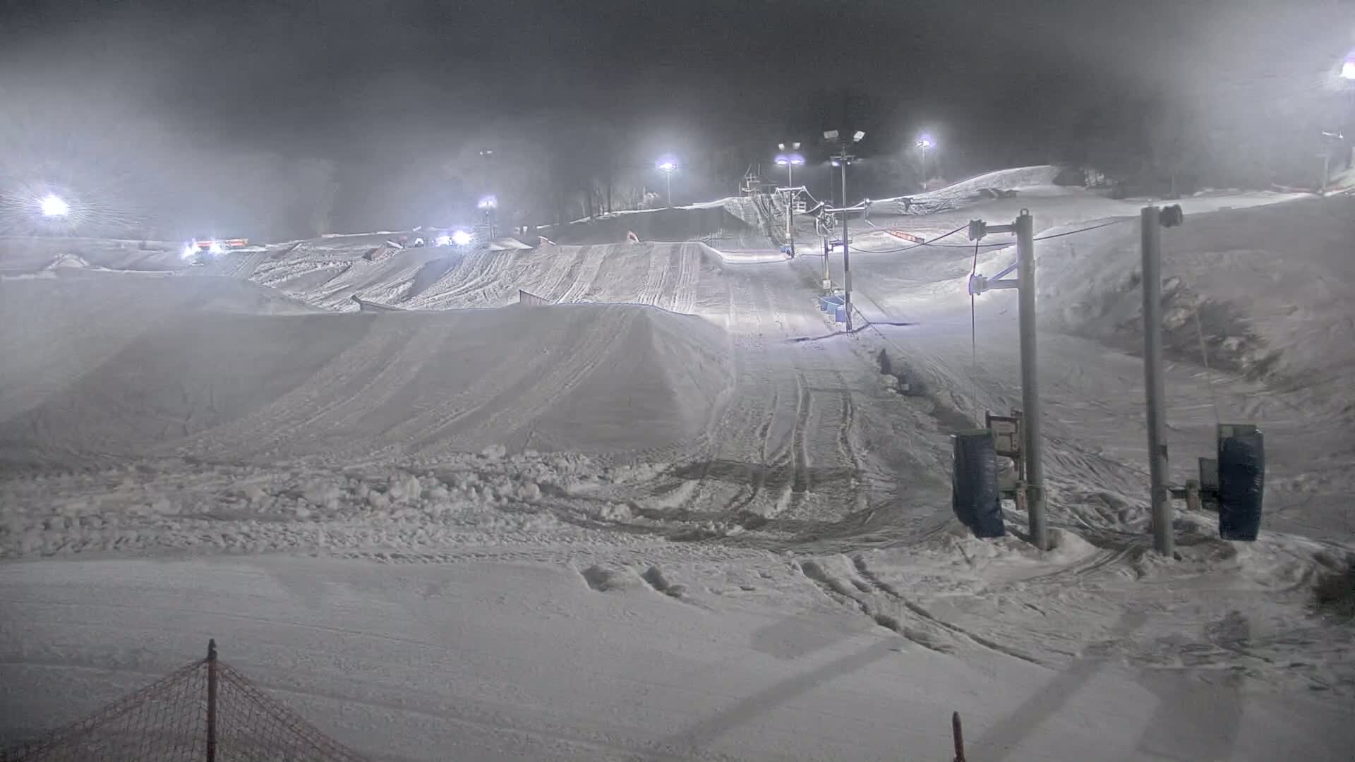 A snow-covered ski slope at night, illuminated by lights, with a ski lift and snow features visible in foggy conditions.