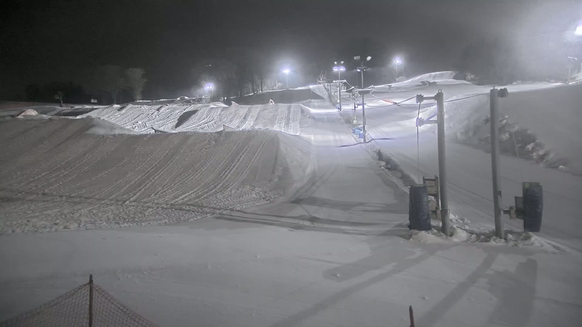 A snow-covered ski slope at night, illuminated by artificial lights, with a ski lift visible in the background under foggy conditions.