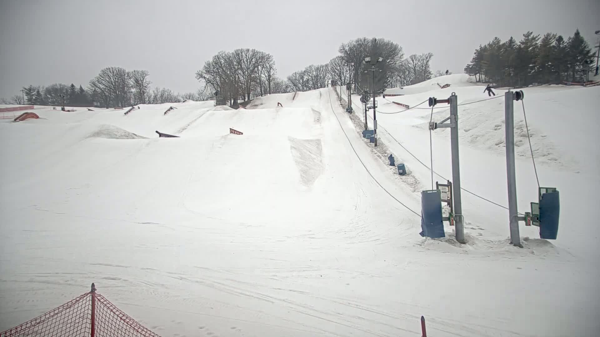 A snow-covered ski slope with a surface lift and several terrain features is visible under an overcast sky.