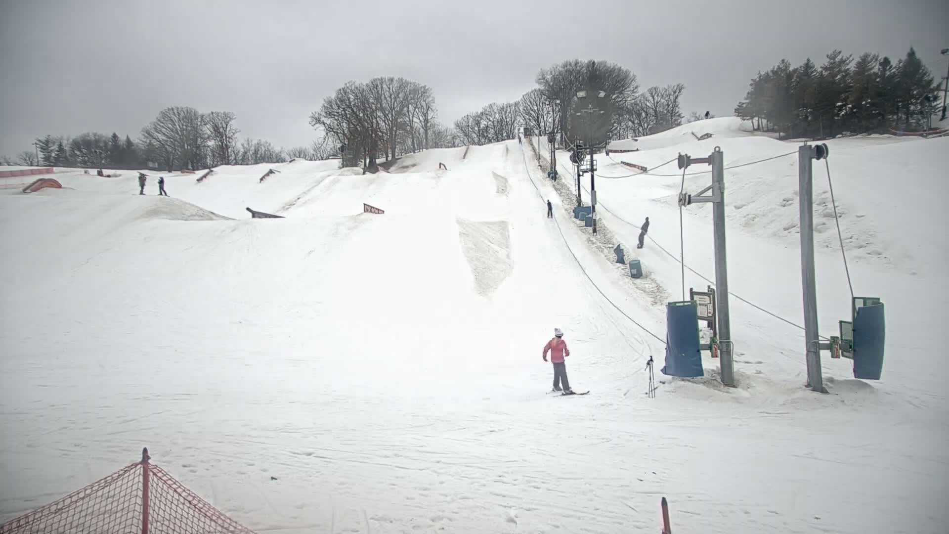 A snowy ski slope with a chairlift and several skiers and snowboarders under an overcast sky.