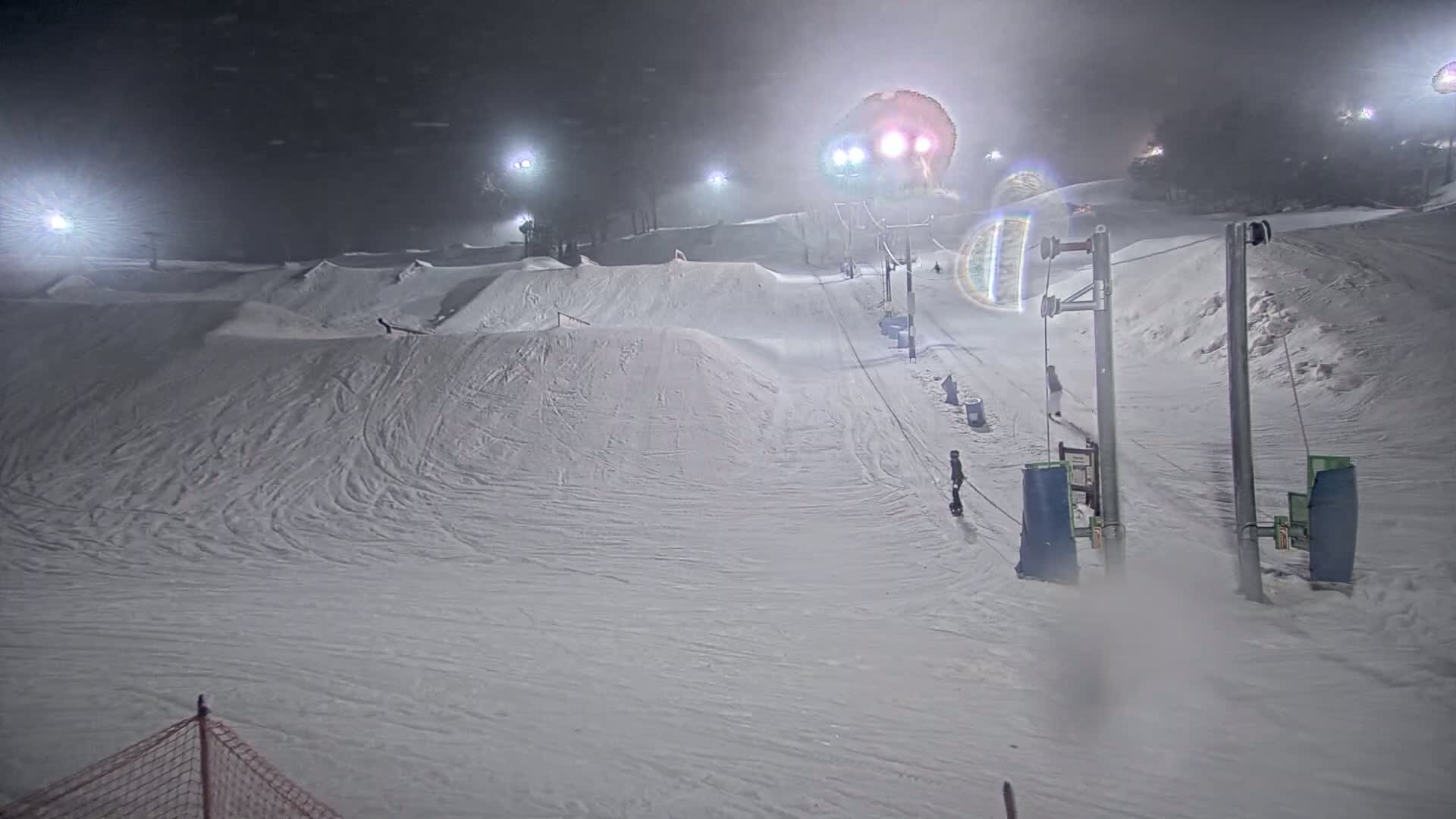 A snow-covered ski slope at night, illuminated by artificial lights, with a ski lift and a few skiers or snowboarders visible in snowy, foggy conditions.