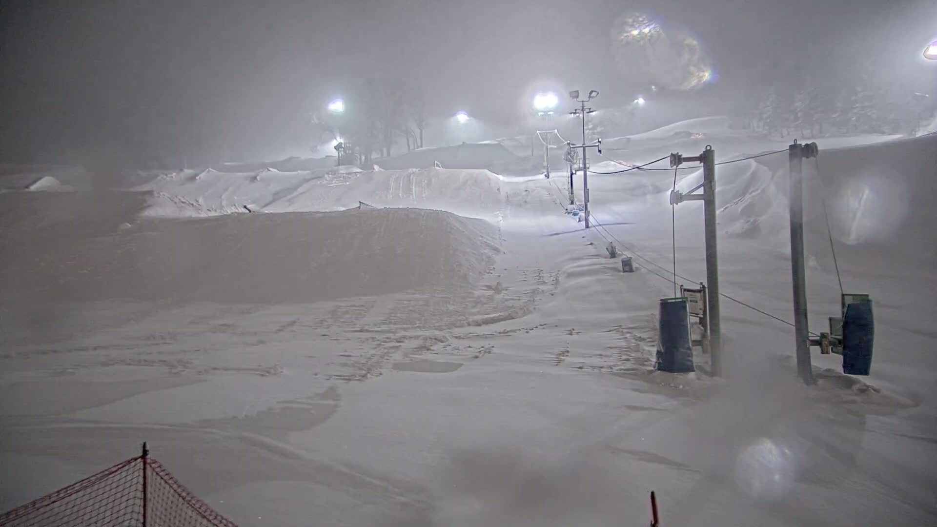 A snow-covered ski slope at night is illuminated by lights, with a surface lift visible in the heavy snowfall.