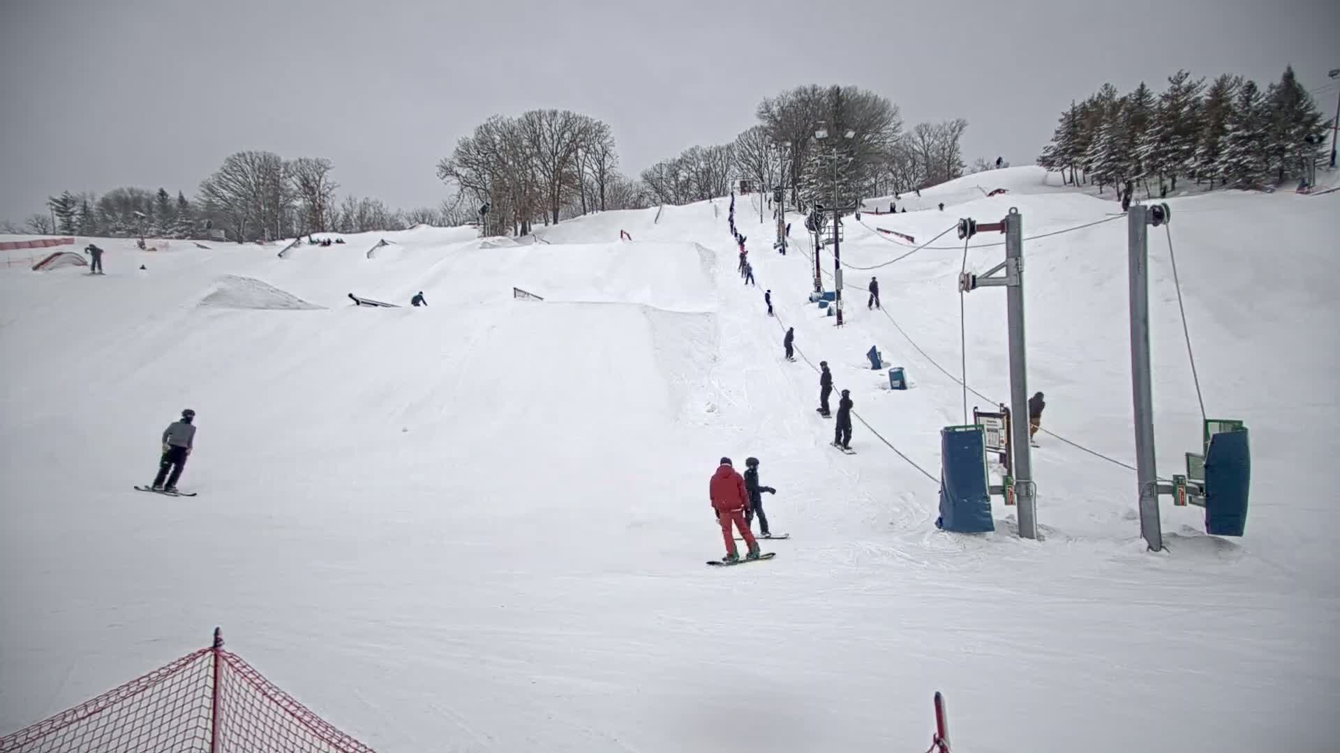 A snow-covered ski hill with several skiers and snowboarders using a surface lift on an overcast day.