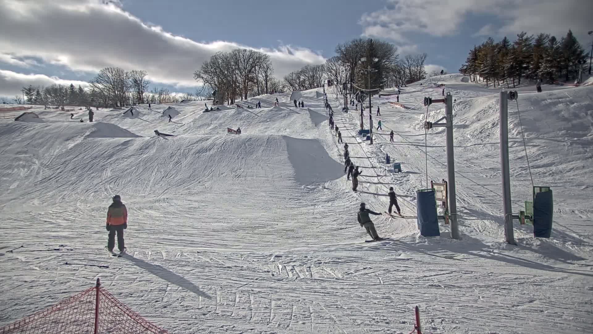 A snow-covered ski hill with several skiers and snowboarders using a surface lift on a partly cloudy day.