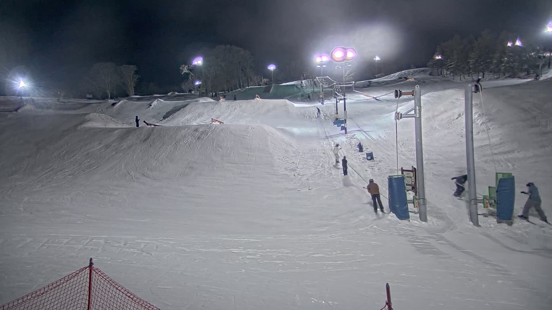 A snow-covered ski slope at night, illuminated by artificial lights, shows several skiers and snowboarders using a surface lift and enjoying terrain features.