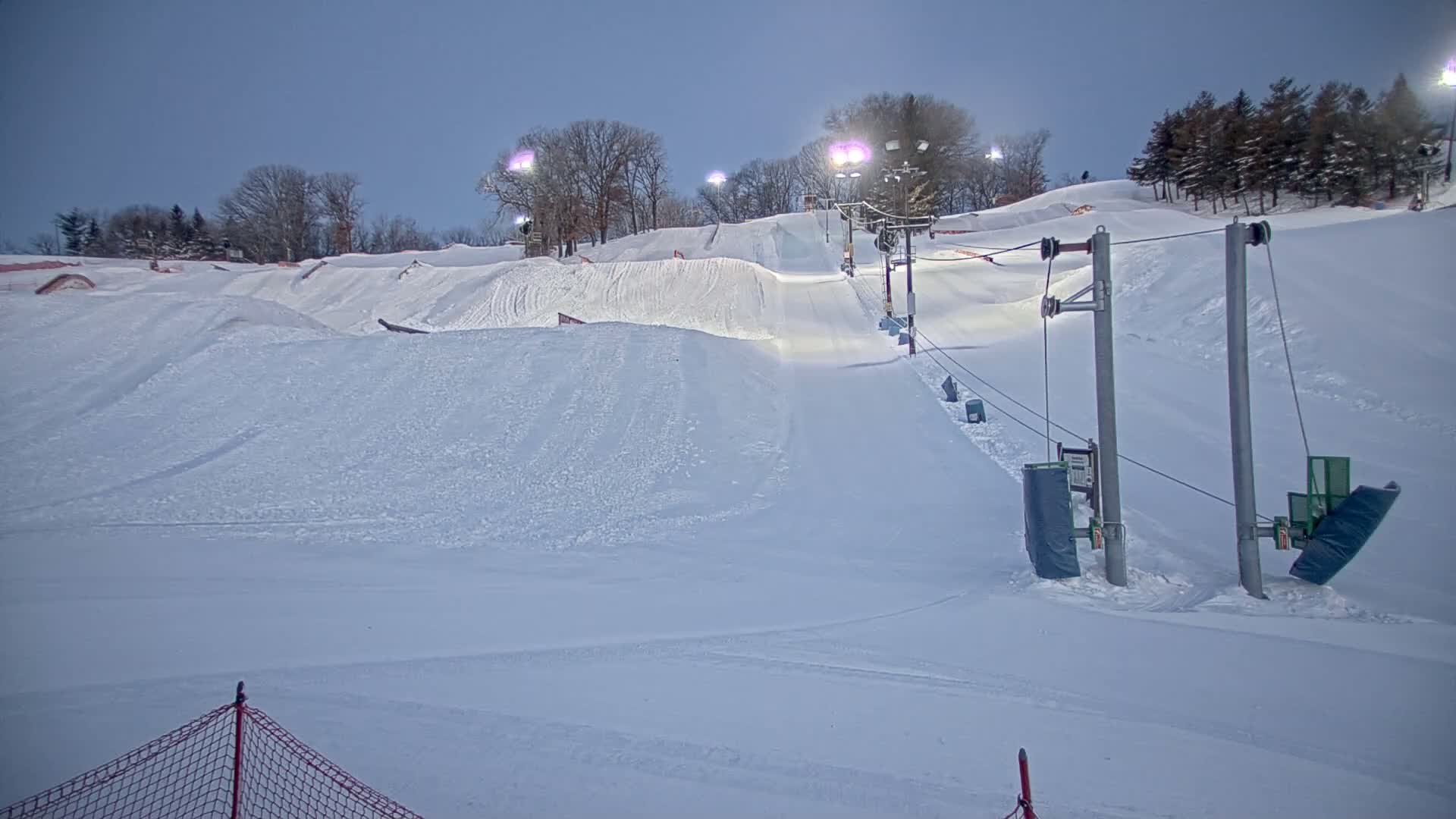 A snow-covered ski hill at night, illuminated by lights, features a ski lift and various snow terrain features.
