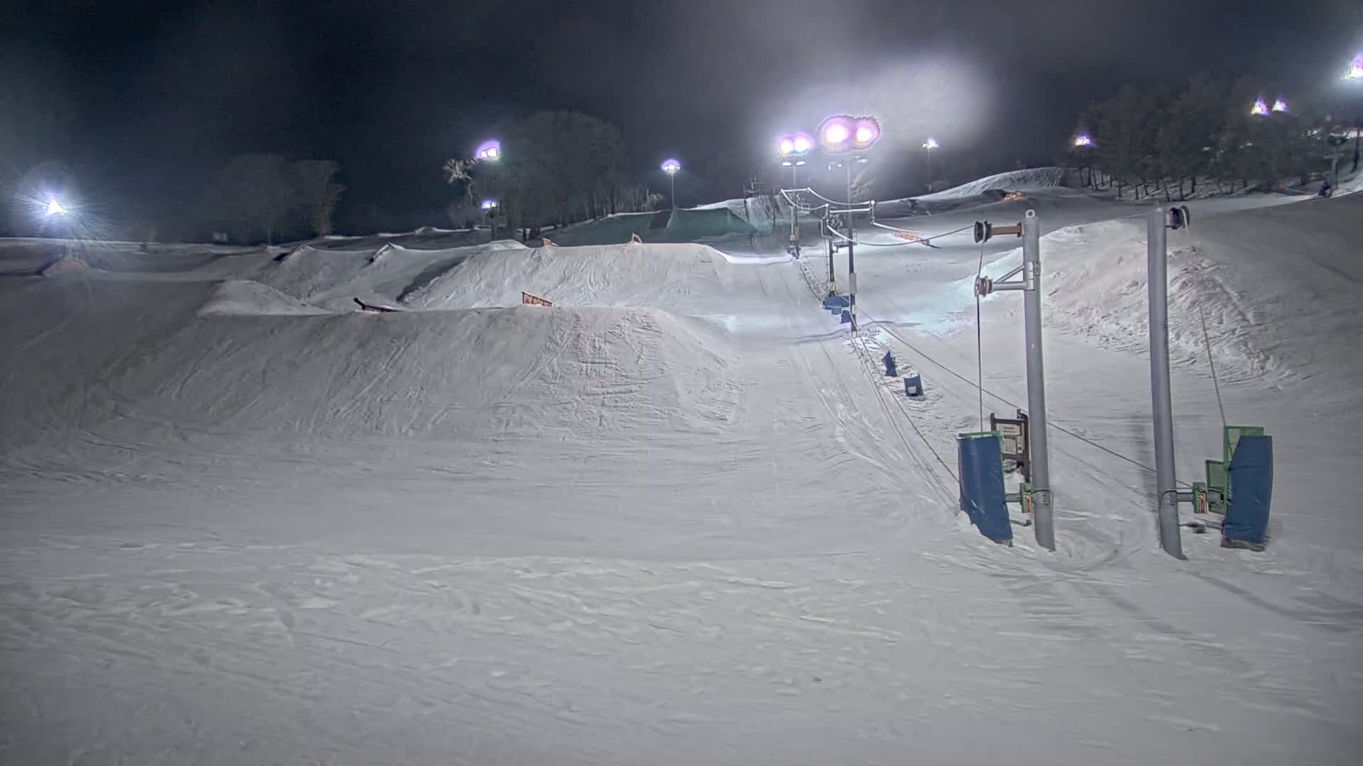 A snow-covered terrain park with a surface lift is illuminated at night.