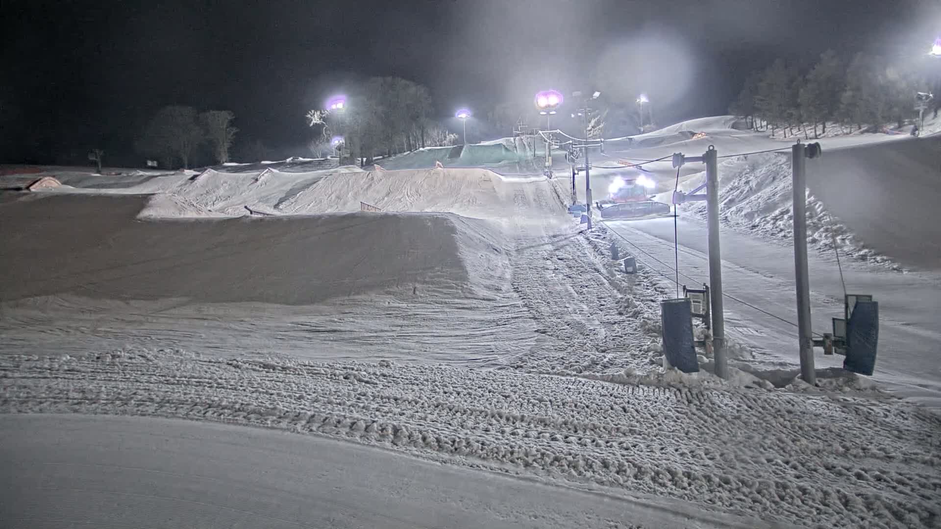 A snowcat grooms a snow-covered ski slope at night, illuminated by artificial lights.