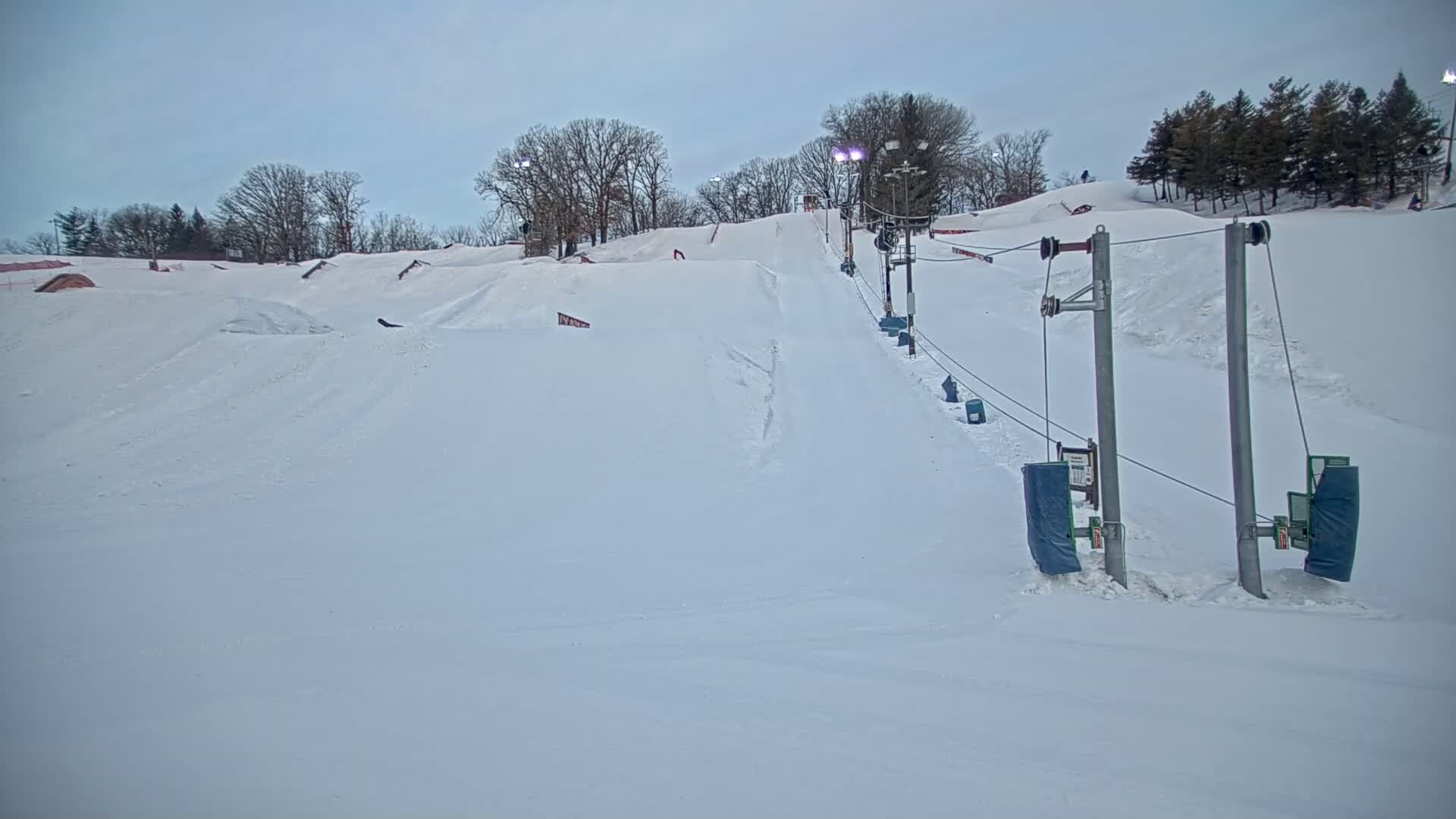 A snow-covered ski slope with a surface lift and terrain park features under a cloudy sky.