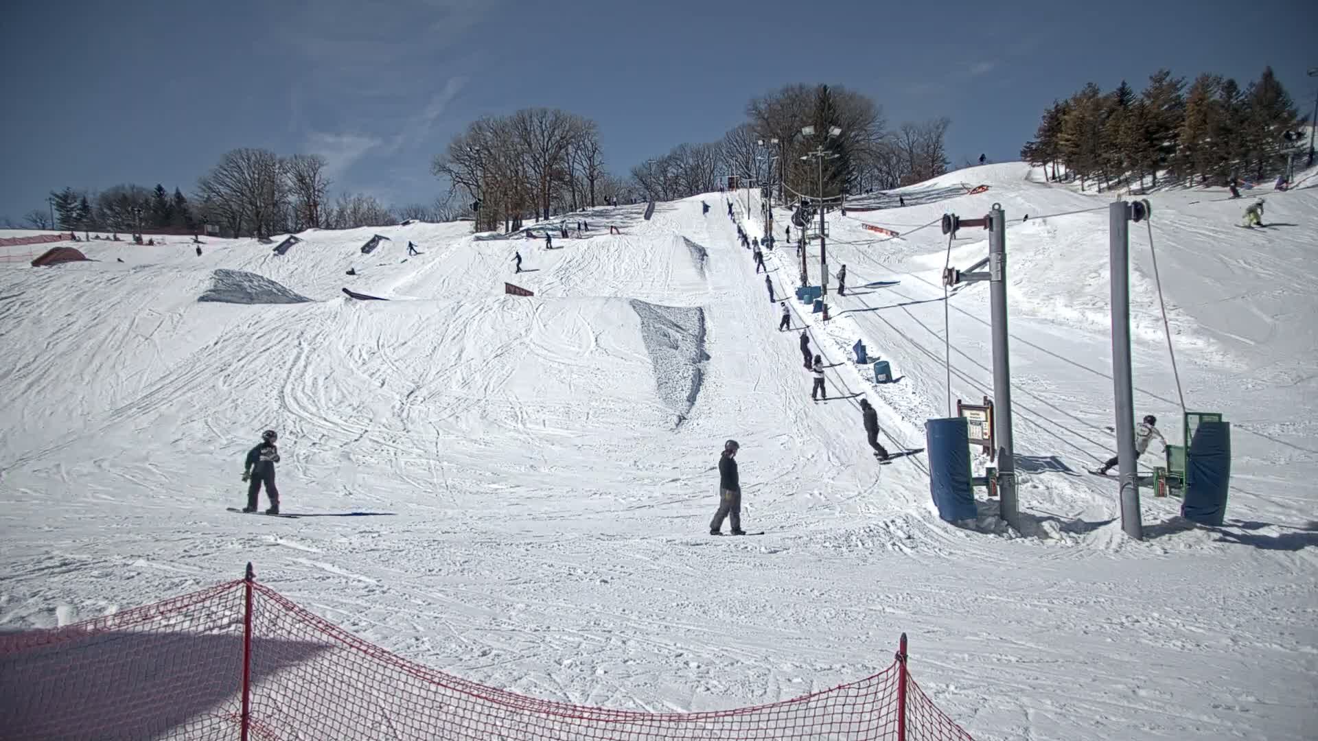 A snowy ski slope with several people snowboarding and skiing under a sunny sky.