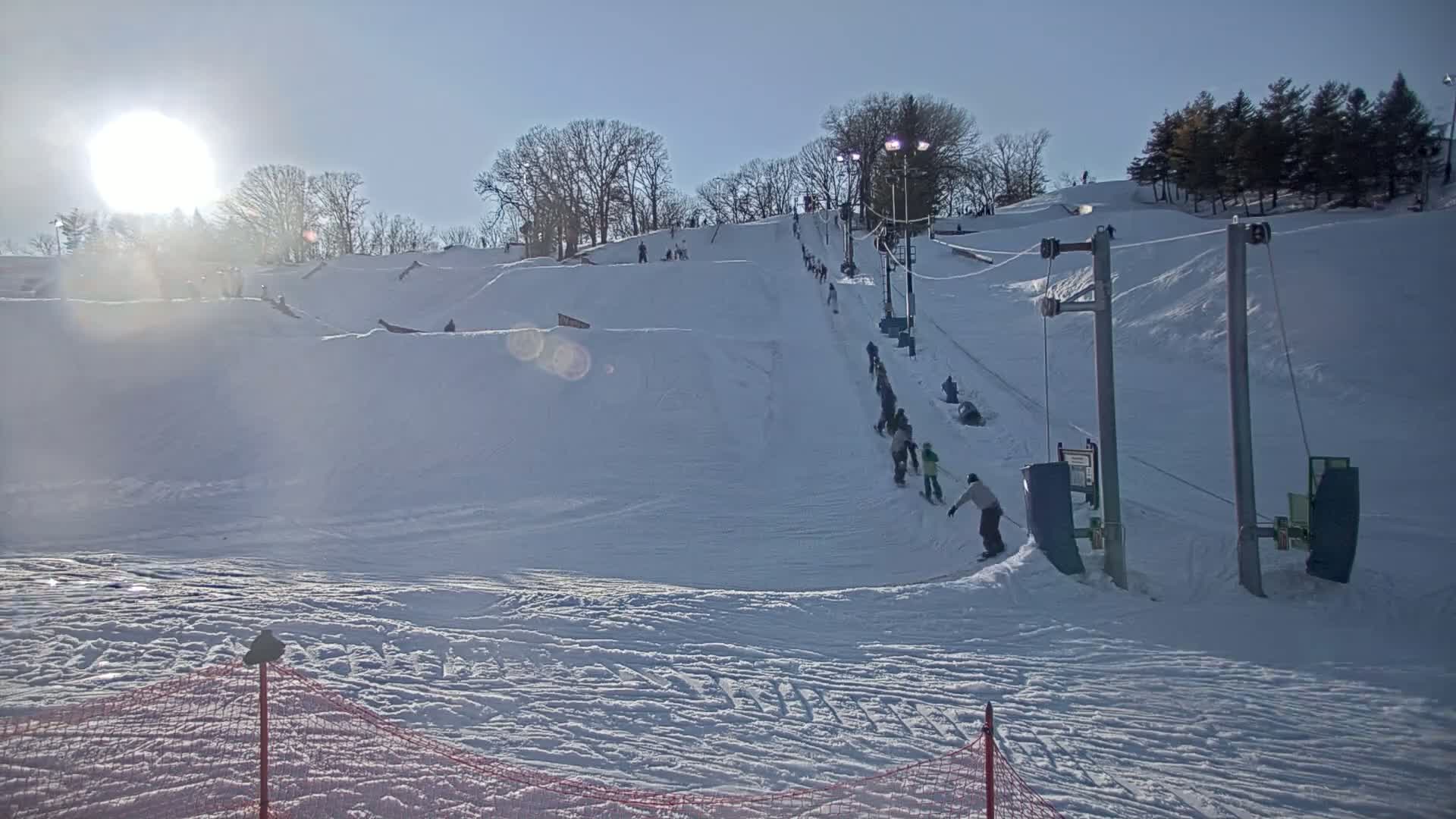 A sunny winter day at a snow-covered ski hill shows a slope with people snowboarding and skiing down toward a ski lift.