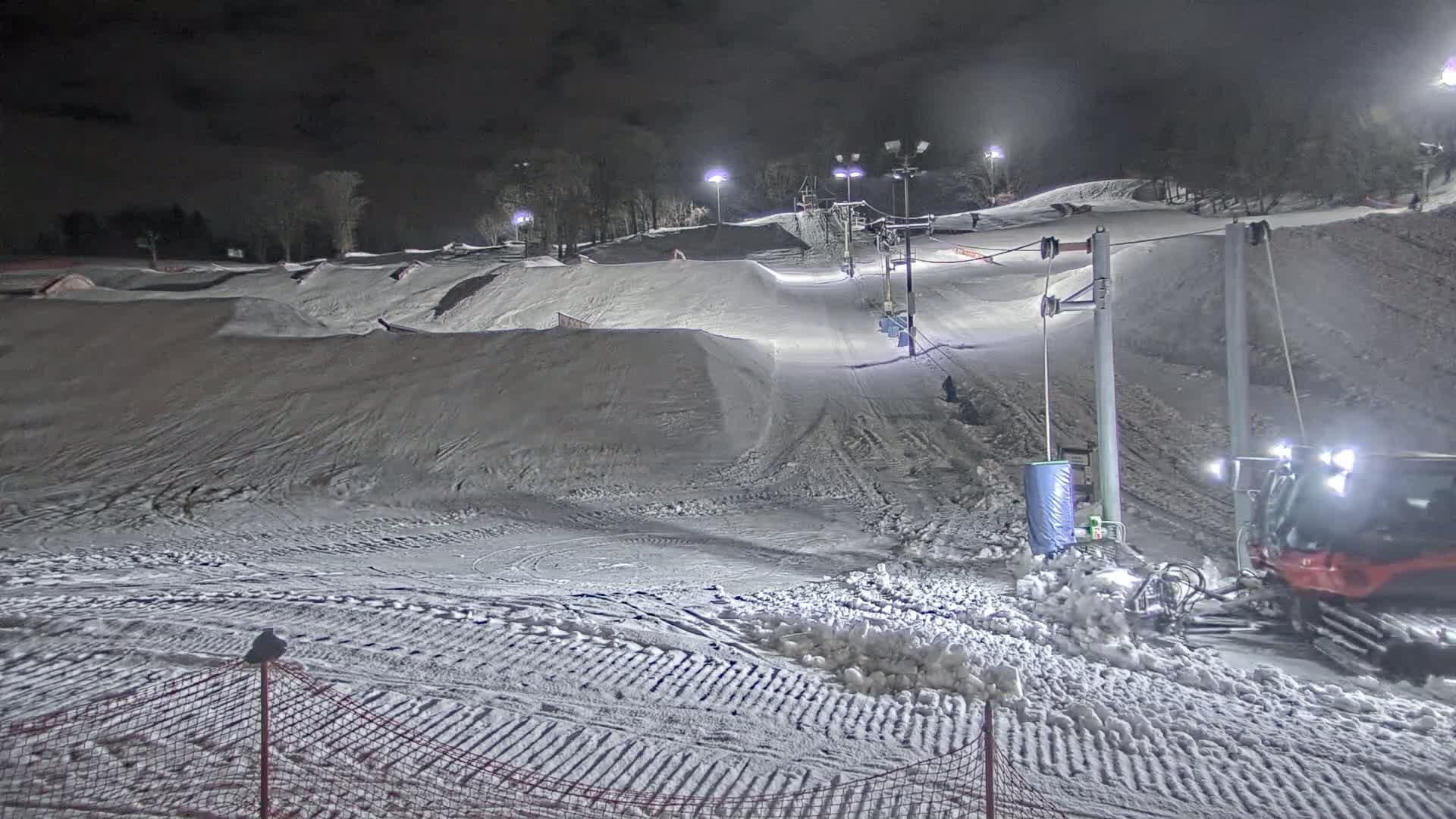 A snow-covered ski slope at night, illuminated by artificial lights, features a ski lift, snow grooming equipment, and various terrain features.