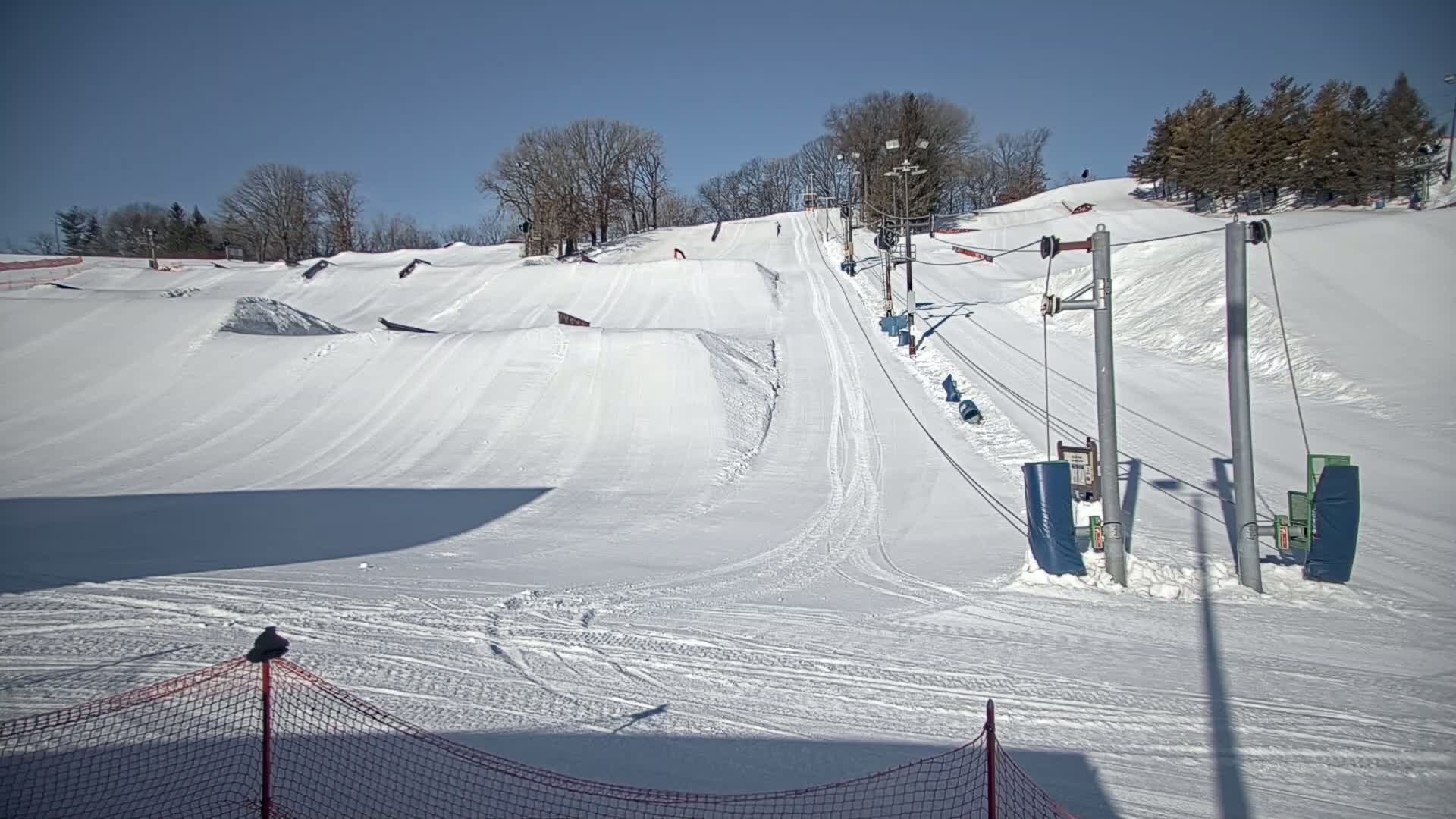 A snowy ski slope with a surface lift and several snow jumps is visible under a clear blue sky.