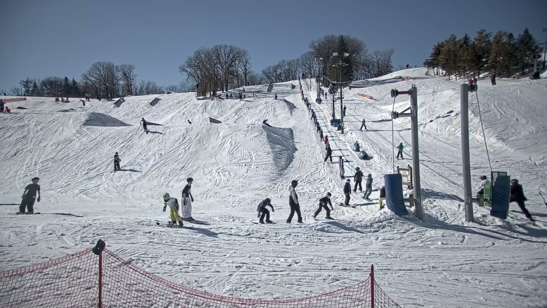 A snow-covered ski hill with several skiers and snowboarders, a ski lift, and terrain features under sunny skies.